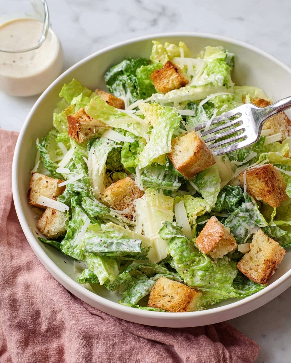 A white bowl filled with fresh green romaine lettuce leaves coated lightly in creamy dressing forms the base layer. On top, golden brown crispy croutons are scattered evenly, adding a crunchy texture. Thin shavings of pale yellow cheese are mixed throughout, peeking between the lettuce and croutons. A silver fork is lifted into the bowl, holding a mix of lettuce and croutons. The bowl sits on a white marbled surface with a soft pink cloth and a small clear glass jug of extra dressing blurred in the background. photo taken with an iphone --ar 4:5 --v 7