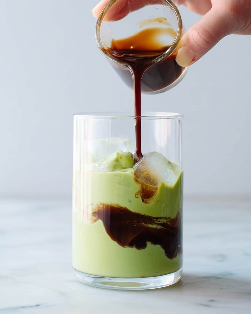 A clear glass cup contains a thick, smooth light green layer that forms the base and mid layers with an icy texture partially visible inside. A woman's hand is pouring a dark brown liquid from a small clear glass cup into the larger cup, creating a swirling pattern where the dark liquid meets the green layer. The scene is set on a white marbled surface with a soft, bright background. photo taken with an iphone --ar 4:5 --v 7