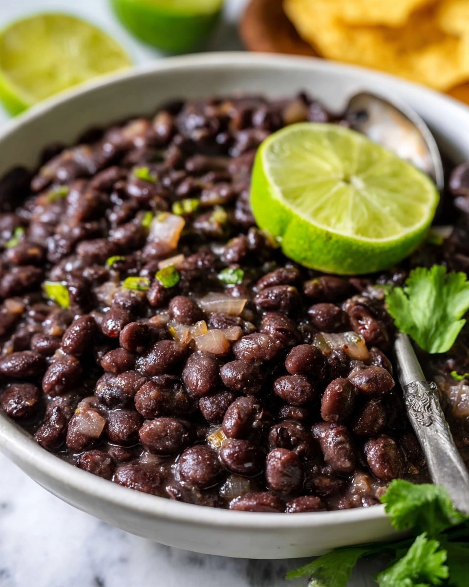 A blue bowl filled with cooked black beans that look soft and shiny, with some beans slightly mashed, topped with two green lime wedges and a sprig of fresh green cilantro on the right side. A light-colored spoon rests inside the bowl, partially covered by the beans. The bowl sits on a white marbled surface scattered with fresh cilantro leaves and lime wedges. In the top right corner, there are yellow tortilla chips and a wooden board holding more lime wedges and a small bunch of cilantro. Photo taken with an iphone --ar 4:5 --v 7