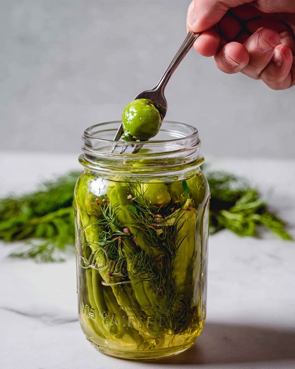 A clear glass mason jar filled with layers of green chili peppers and delicate yellow-green dill sprigs immersed in a light yellowish-green liquid, with a single dark red dried chili pepper visible near the top floating among the herbs and peppers. The jar sits on a white marbled surface with a few green chili peppers scattered around. The jar's embossed label is slightly visible, and the image has soft lighting that highlights the fresh and natural textures of the ingredients photo taken with an iphone --ar 4:5 --v 7