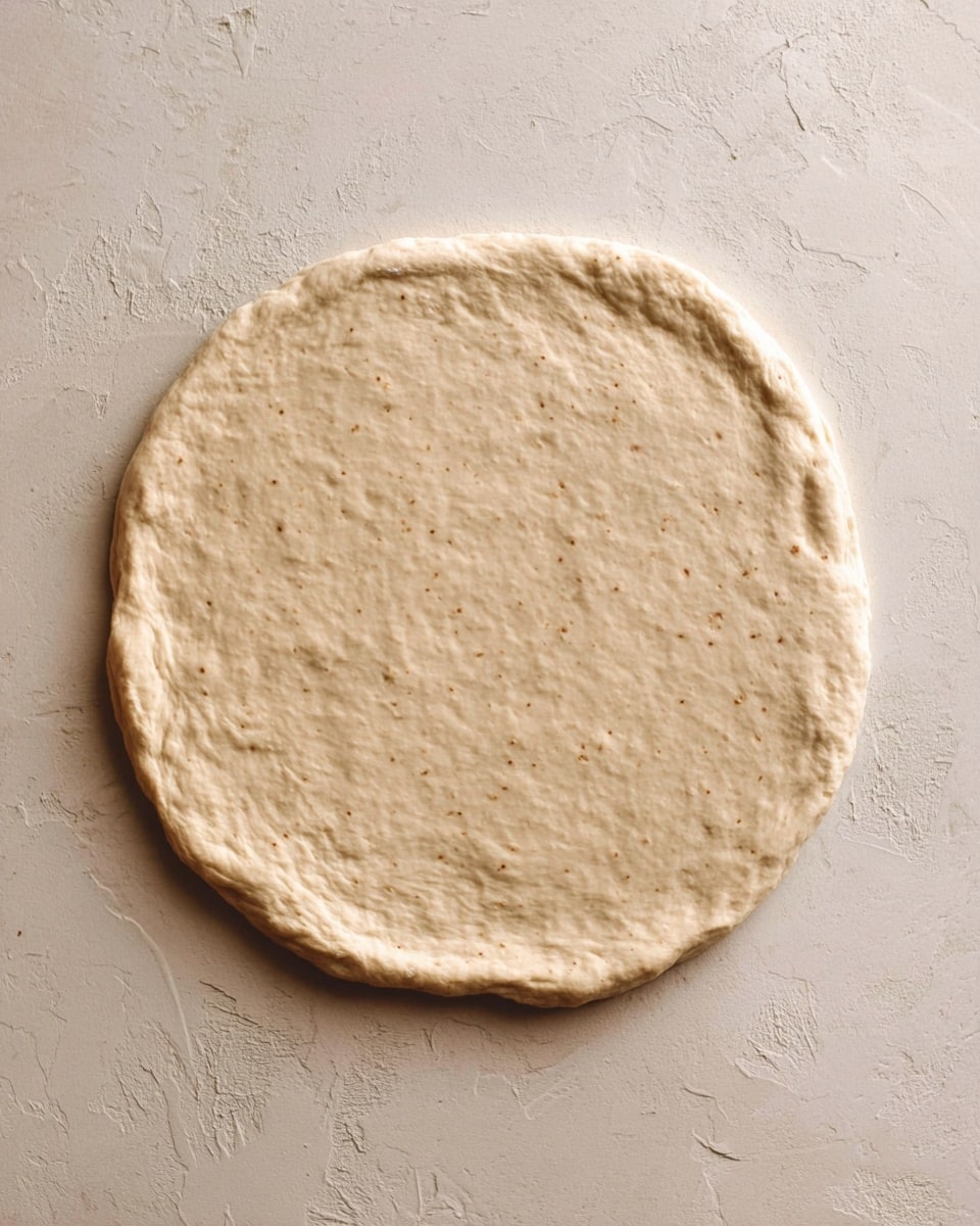 A single, round dough layer with a slightly uneven edge lies flat on a textured white marbled surface. The dough is light beige with small specks and has a soft, slightly puffy texture showing gentle creases and dimples across the surface. It appears uncooked and smooth, taking up most of the frame. Photo taken with an iphone --ar 4:5 --v 7