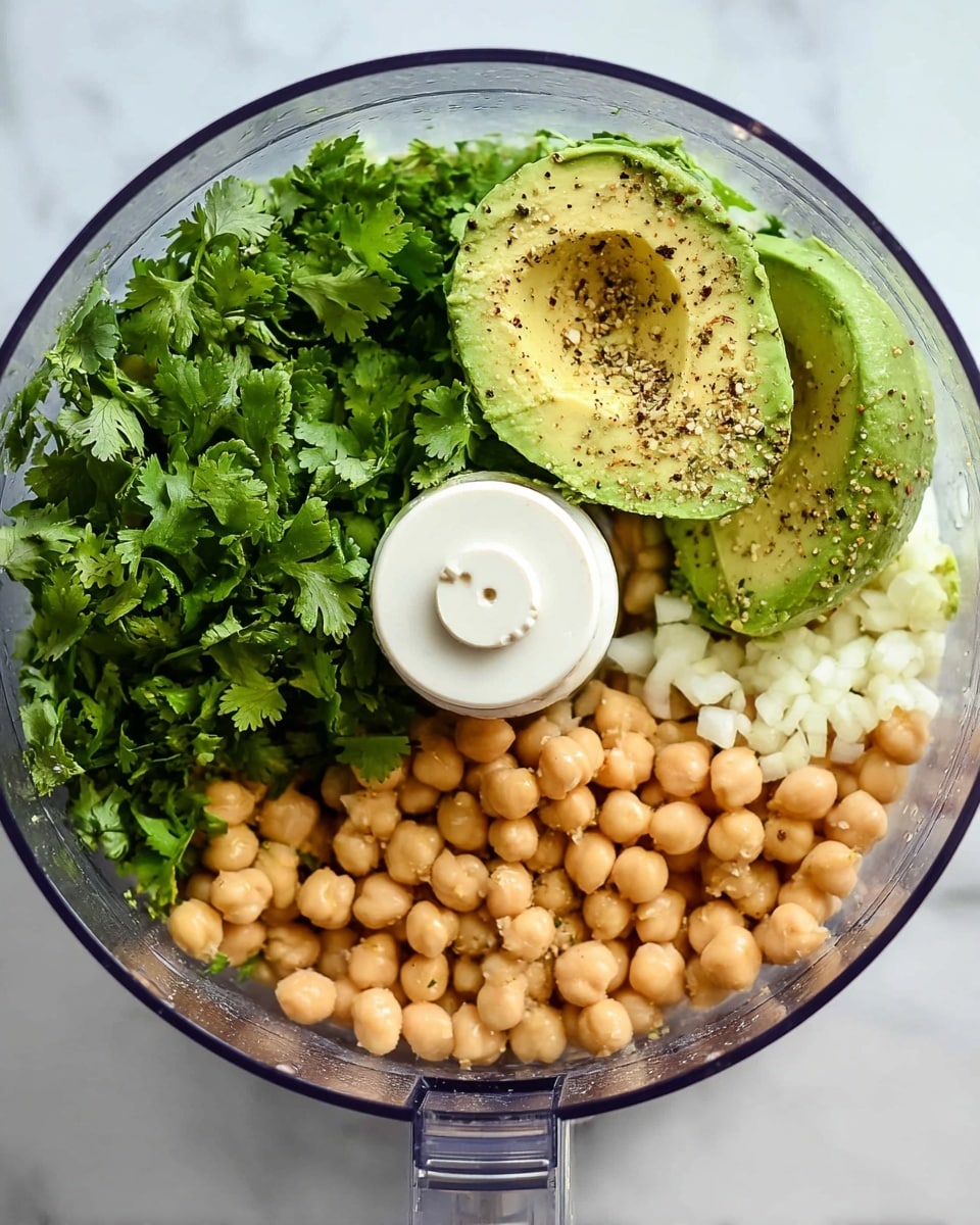 The image shows the inside of a clear food processor bowl filled with fresh ingredients. At the bottom right, there is a layer of light tan chickpeas, slightly round. On the left side, there is a bunch of bright green cilantro leaves with ruffled edges. Above the chickpeas near the center-right, two halves of a green avocado with smooth texture and a hollow center are visible, sprinkled with black pepper. Small white chopped garlic pieces sit on the chickpeas near the cilantro. The food processor’s white plastic blade attachment is visible in the middle. The whole setup is on a white marbled surface. photo taken with an iphone --ar 4:5 --v 7