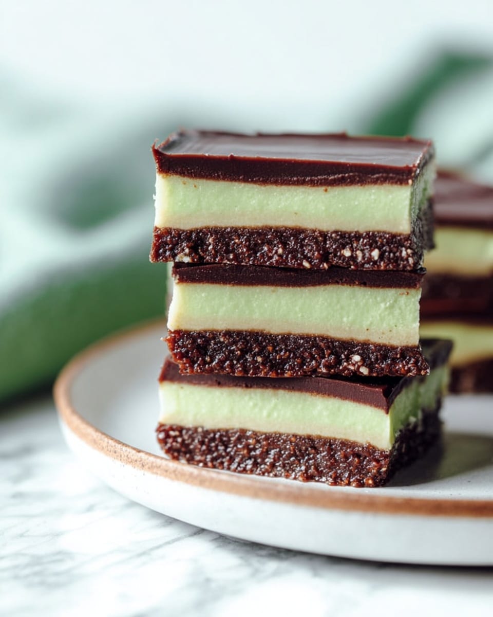The image shows a close-up of four stacked dessert bars on a white plate with a thin brown rim, placed on a white marbled surface. Each bar has three layers: the bottom layer is dark brown and textured, looking like a dense chocolate base; the middle layer is thick, creamy, and pale green; the top layer is a smooth, thin dark brown chocolate layer. The bars are arranged so the layers are clearly visible from the side, and the background is softly blurred with light colors, including a greenish shape and some blue fabric. Photo taken with an iphone --ar 4:5 --v 7