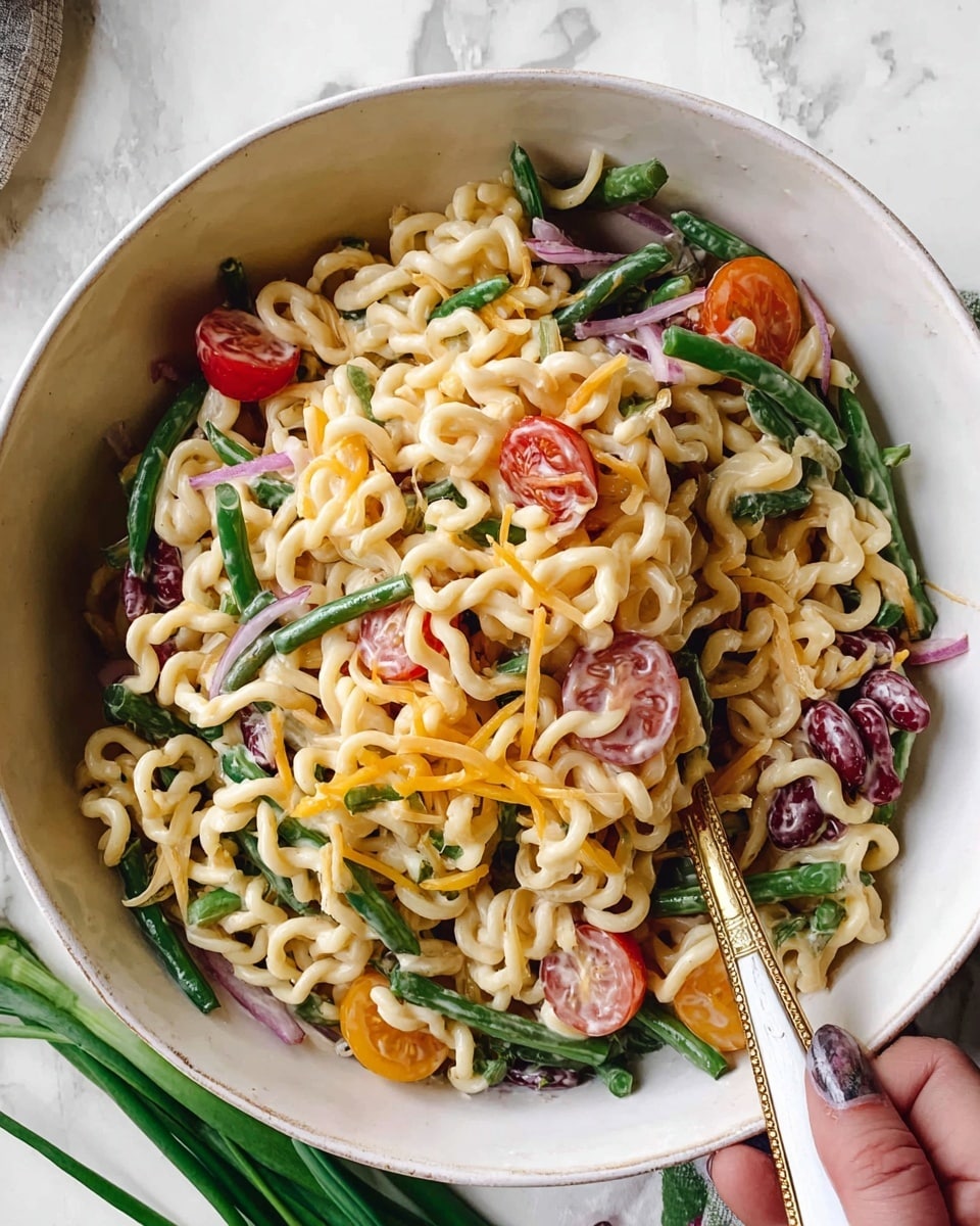 A large white bowl holds a mixed pasta salad with three main layers: curly cooked noodles form the base, creamy dressing coats the noodles and vegetables, and fresh green beans, halved cherry tomatoes, shredded yellow cheese, thinly sliced red onions, and kidney beans are scattered throughout, adding red, green, and purple colors. A silver fork with a white and gold handle is partly buried in the salad, and a woman's hand is slightly visible holding the bowl or utensils. The bowl rests on a white marbled surface with some green onions partially visible at the edge. photo taken with an iphone --ar 4:5 --v 7