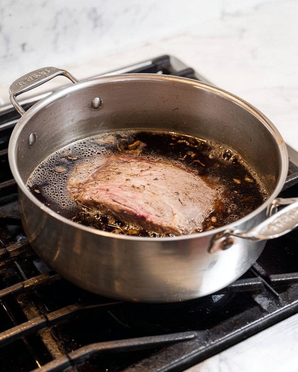 A large silver pot sits on a stove with dark liquid inside, in which a thick piece of meat is cooking. The meat is mostly brown but has a pinkish center showing it is partly cooked. The liquid has bubbles and some foam on the surface, with bits of seasoning floating around. The pot has handles on both sides and rests on black stove grates. The background is a white marbled texture photo taken with an iphone --ar 4:5 --v 7