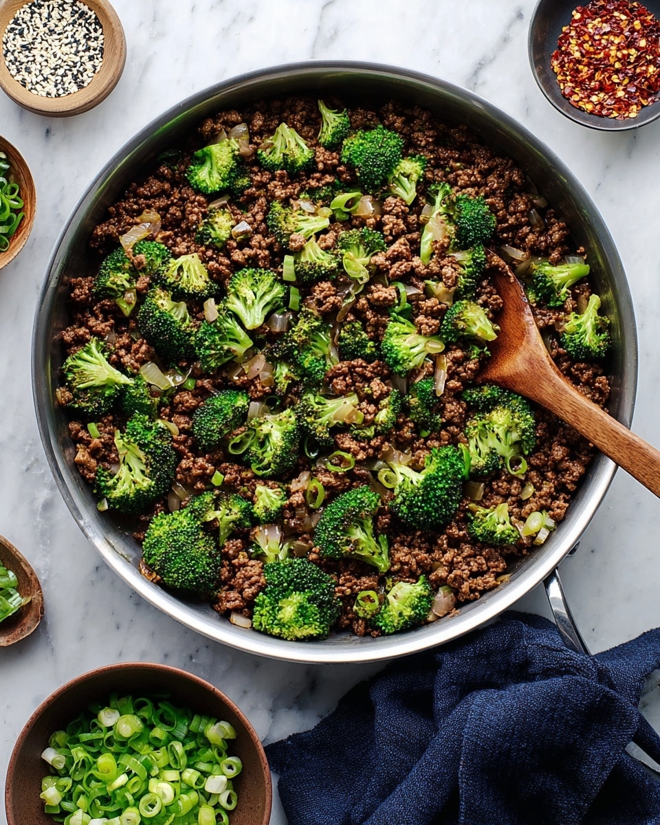 A large silver frying pan holds a dish made of dark brown cooked ground meat mixed with small pieces of onion and bright green broccoli florets spread evenly on top. The broccoli adds a fresh look with its crisp texture and vibrant color contrasting with the rich brown meat. A wooden spoon rests inside the pan on the right side, angled slightly upward. The pan is on a white marbled surface, surrounded by small bowls: one with red chili flakes, one with white and black sesame seeds, and a brown bowl filled with chopped green onions. A dark blue cloth sits underneath the pan handle. Photo taken with an iphone --ar 4:5 --v 7