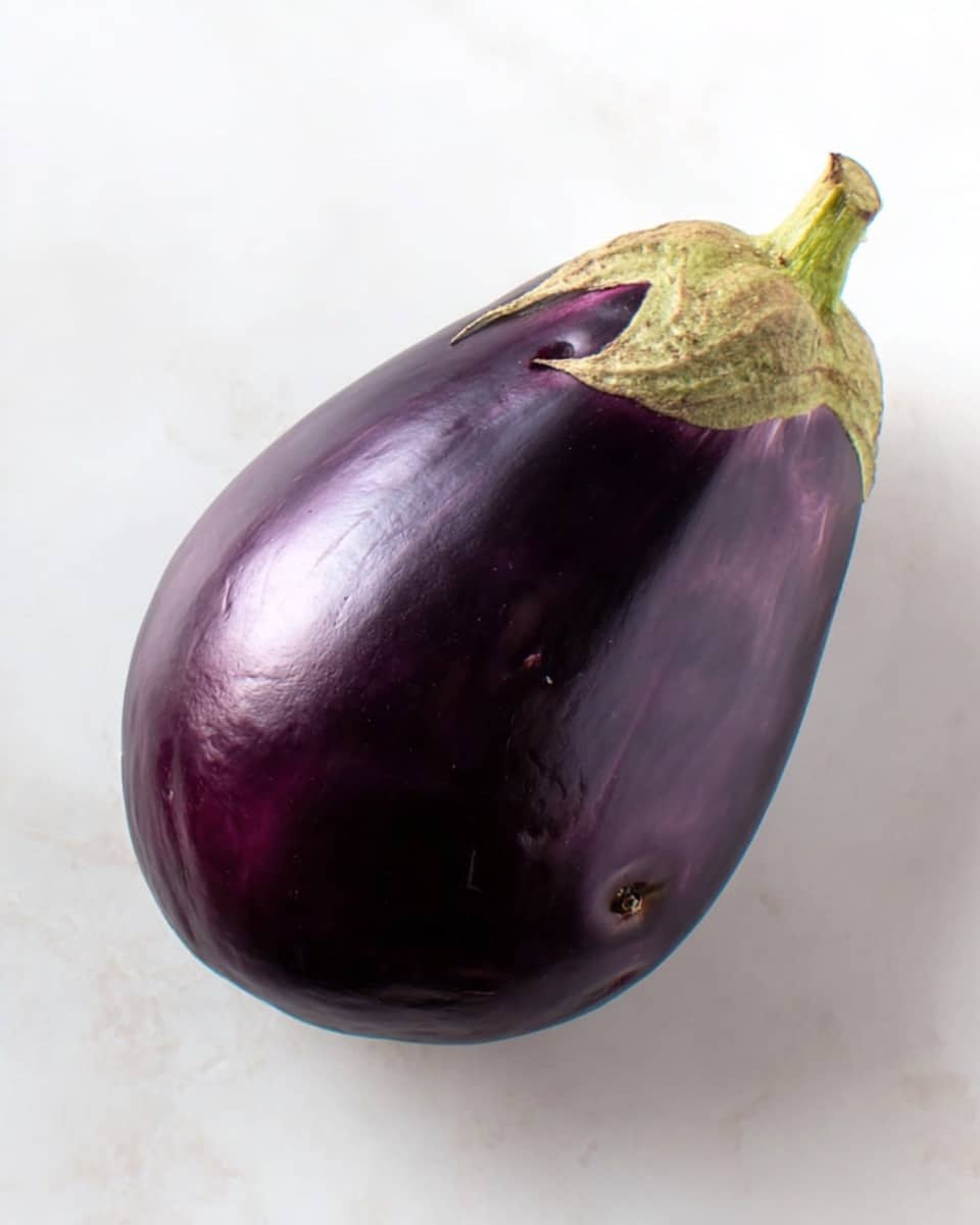 The image shows a whole eggplant placed on a white marbled surface. The eggplant has a smooth, shiny deep purple skin with a few small natural marks. It has a light green stem attached at the top. The shape of the eggplant is round and slightly wide at the lower half, tapering slightly towards the stem. The lighting creates mild reflections on the eggplant’s surface, highlighting its smooth texture. Photo taken with an iphone --ar 4:5 --v 7