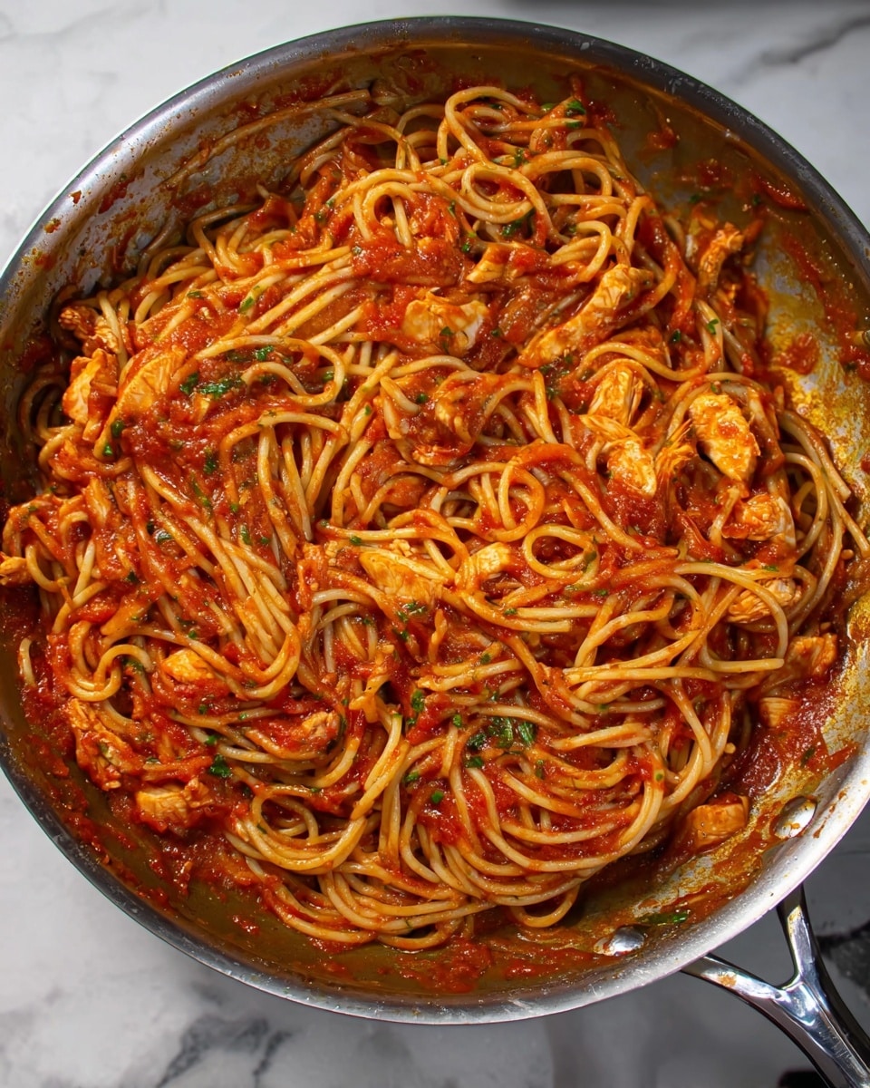 In a silver pan, there is a serving of spaghetti mixed with a thick red tomato sauce. The sauce is spread all over the noodles and has small pieces of cooked chicken and green herbs visible throughout. The noodles are long and lightly coated by the sauce, with some tangled in the center. The pan is placed on a white marbled surface, and the shiny steel handle is visible, showing some sauce stains near its base. The overall look is warm and hearty with a mix of red, yellow, and green colors. photo taken with an iphone --ar 4:5 --v 7