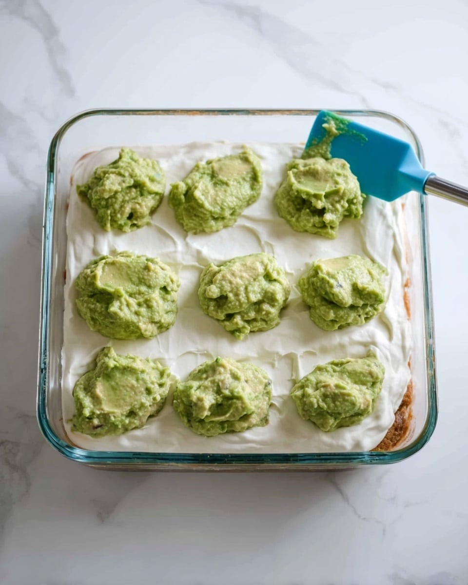 A clear glass rectangular dish contains a layered food item. The bottom layer looks light brown and slightly moist. The second layer is thick and smooth, creamy white, covering the entire dish evenly. On top of this white layer, there are eight dollops of a chunky green mixture that looks like mashed avocado, spread unevenly but mostly centered in rows. A blue spatula rests on the top right corner of the dish, touching the green mixture. The dish is placed on a white marbled surface. photo taken with an iphone --ar 4:5 --v 7