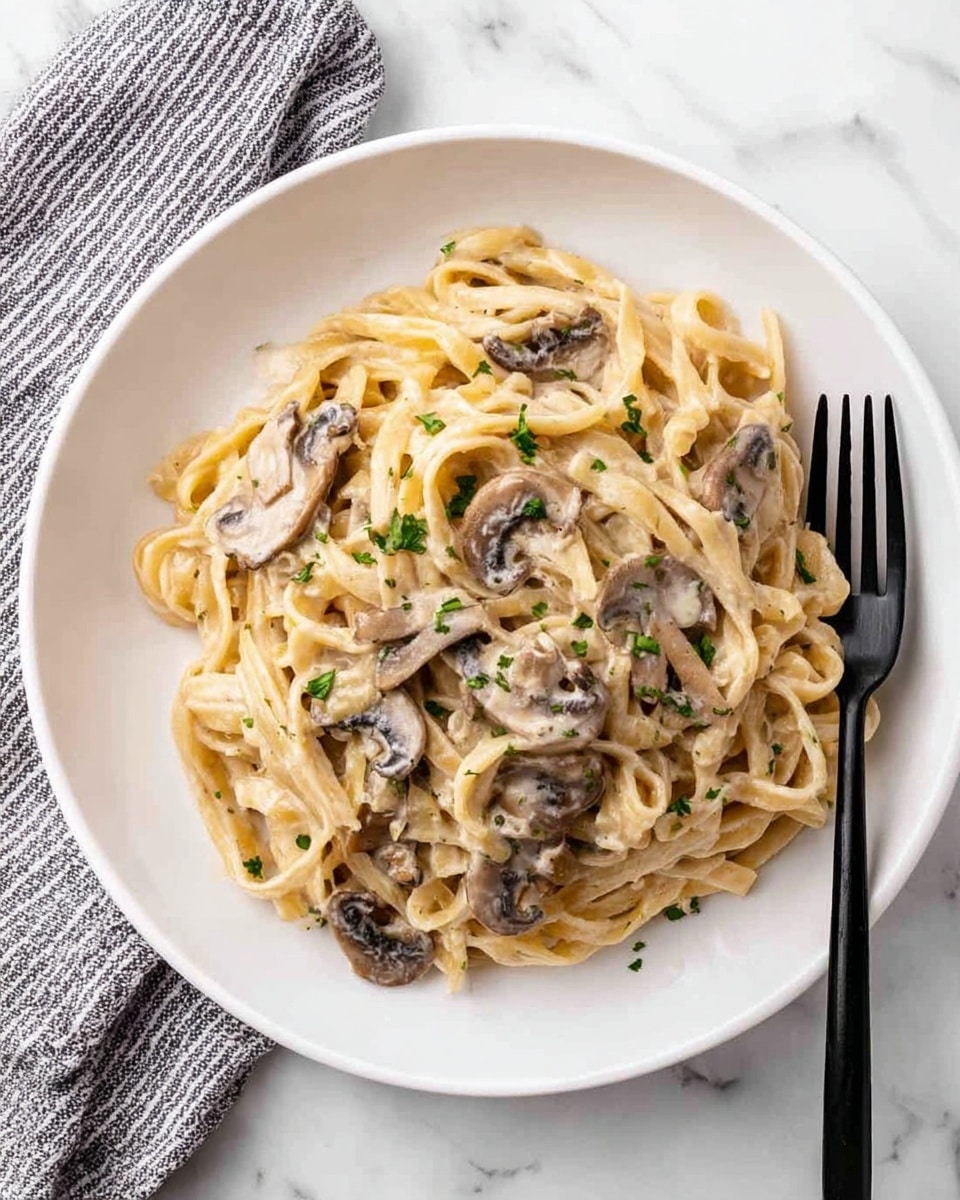 The image shows a white plate filled with creamy pasta mixed with sliced mushrooms. The pasta is pale beige and covers most of the plate, with the mushrooms scattered on top and beneath the noodles, showing a dark brown color and soft texture. The creamy sauce gives a smooth, slightly shiny look to the pasta, while some small green herb bits, likely parsley, are sprinkled over the dish for color contrast. A black fork rests on the right edge of the plate. The plate is placed on a white marbled surface with a gray and white striped cloth napkin to the left. Photo taken with an iphone --ar 4:5 --v 7