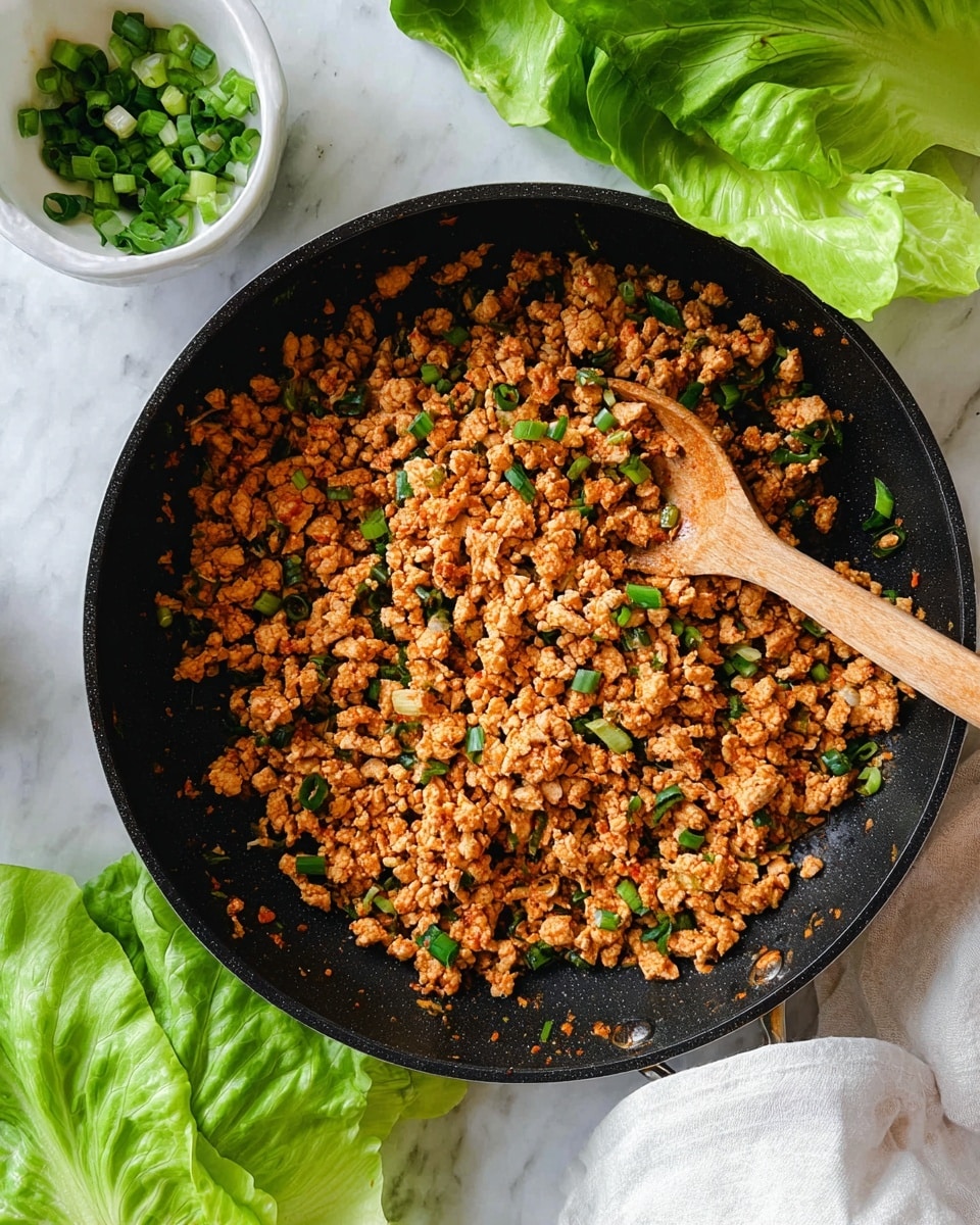 A black pan filled with cooked ground chicken mixed with small pieces of green onion and finely chopped vegetables, showing an orange-brown color from spices and sauce, with a wooden spoon resting inside the pan. Around the pan, there are fresh green lettuce leaves on a white marbled surface, and a small white bowl filled with chopped green onions near the bottom left corner. Photo taken with an iphone --ar 4:5 --v 7