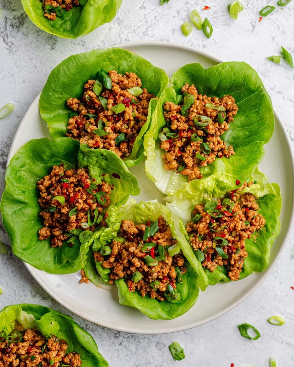 A white plate with five large green lettuce leaves arranged in a circle, each leaf holding a layer of finely chopped, cooked meat mixed with small bits of green onions and red chili flakes, giving the meat a slightly shiny and textured look, some meat pieces scattered slightly outside the lettuce leaves. Around the plate, there are small pieces of green onion scattered on a white marbled surface. photo taken with an iphone --ar 4:5 --v 7