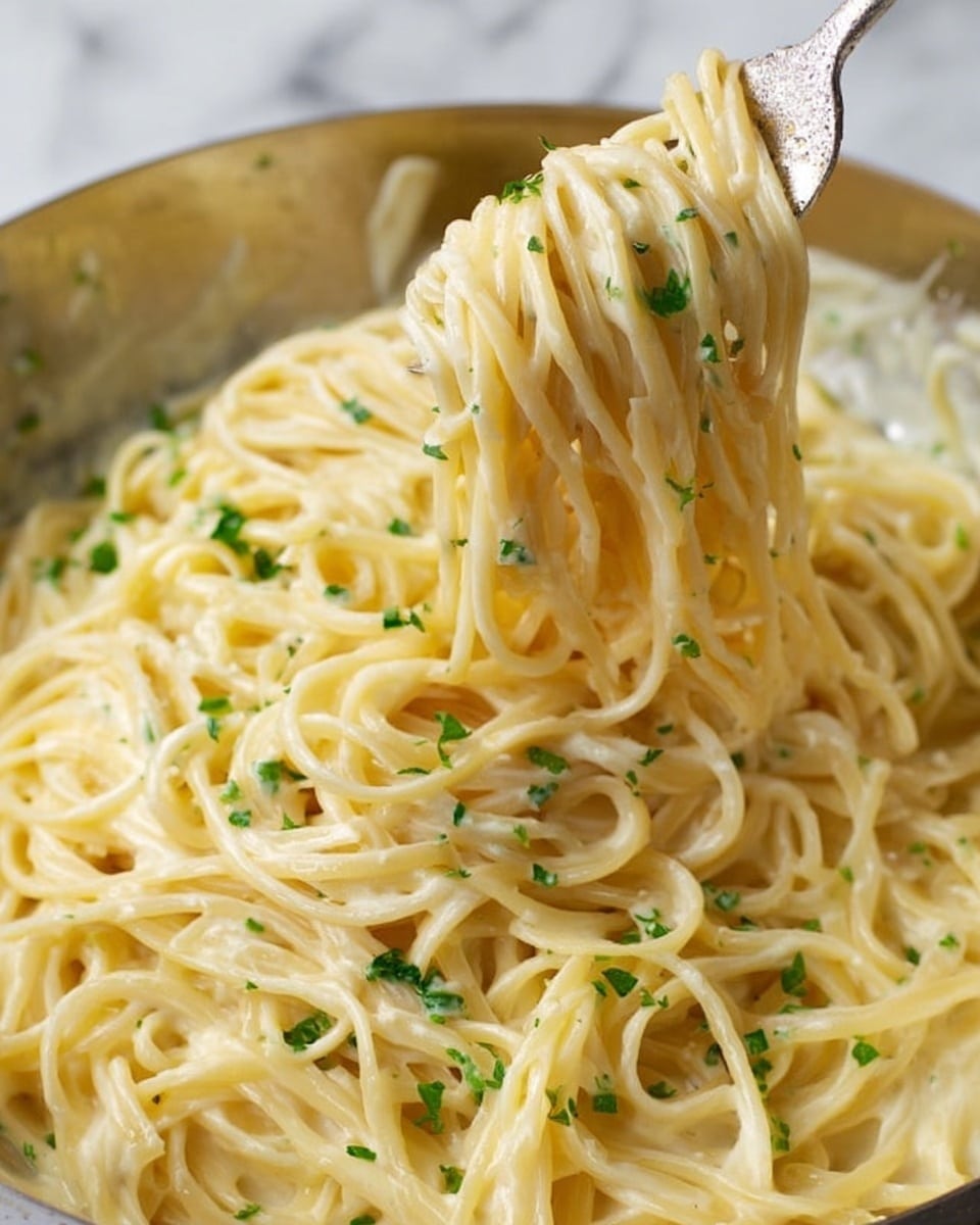 A close-up view of creamy spaghetti noodles covered with light yellow cheese sauce, mixed with small bits of green herbs sprinkled evenly on top. The spaghetti strands are thick and smooth, tangled together while being lifted by a fork. The dish is in a reflective metal bowl showing the creamy texture of the sauce clinging to the pasta. The background is a white marbled surface. Photo taken with an iphone --ar 4:5 --v 7