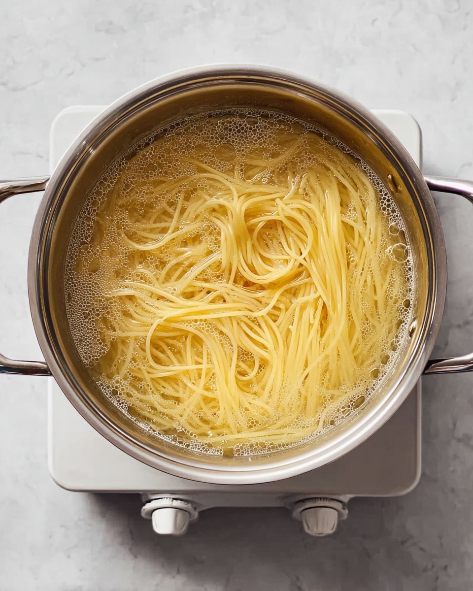 A top view of a silver pot filled with thin yellow spaghetti noodles boiling in clear water with small bubbles on the surface. The pot has two handles and sits on a white electric stove knob, all placed on a white marbled background. Photo taken with an iphone --ar 4:5 --v 7