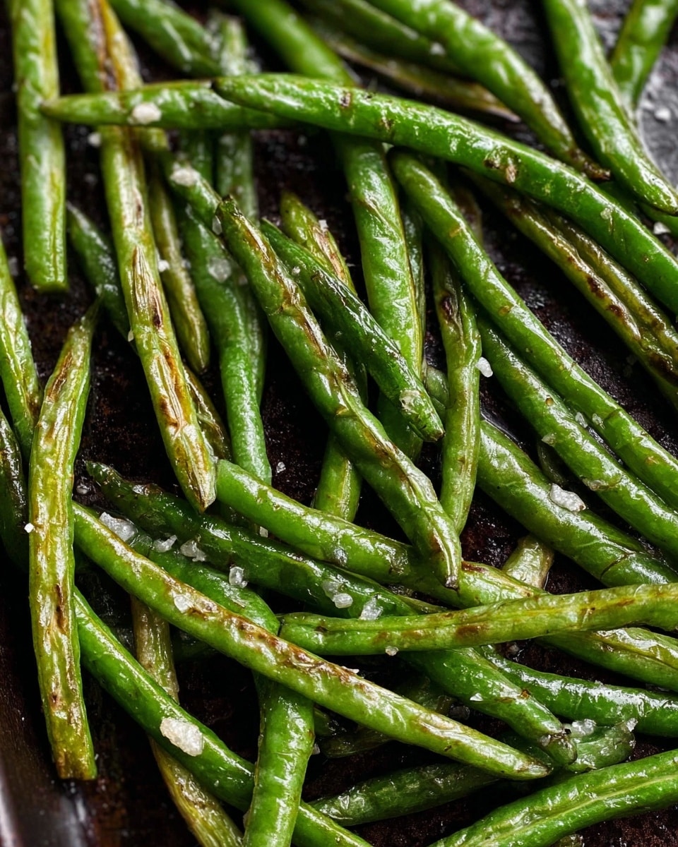 The image shows many cooked green beans spread out on a dark pan. The green beans are shiny, slightly wrinkled, and have light browning in some areas, showing they have been roasted or cooked. Small bits of white seasoning are scattered over the green beans, adding texture. The green beans are arranged in a loose, natural way, filling the frame with their bright green color and glossy surface. The background of the image is a white marbled texture. photo taken with an iphone --ar 4:5 --v 7