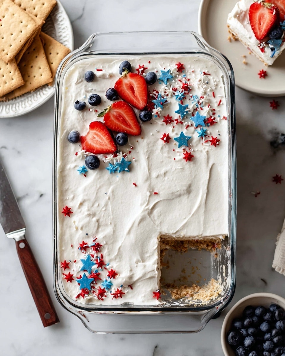 This image shows a clear glass rectangular dish filled with a layered dessert. The top layer is thick, smooth white cream with subtle vanilla specks, spread evenly across the surface. On top, there are three strawberry halves placed in a line down the middle, surrounded by some whole blueberries and small red, white, and blue star-shaped sprinkles scattered around them. One piece is missing from the bottom right corner, showing layers beneath the cream, including a light brown crumbly layer and some red berry pieces. The dish sits on a white marbled surface with three graham crackers to the left and a white plate with a slice of the dessert and blueberries to the upper right. A knife with a wooden handle is partly visible on the left side. Photo taken with an iphone --ar 4:5 --v 7