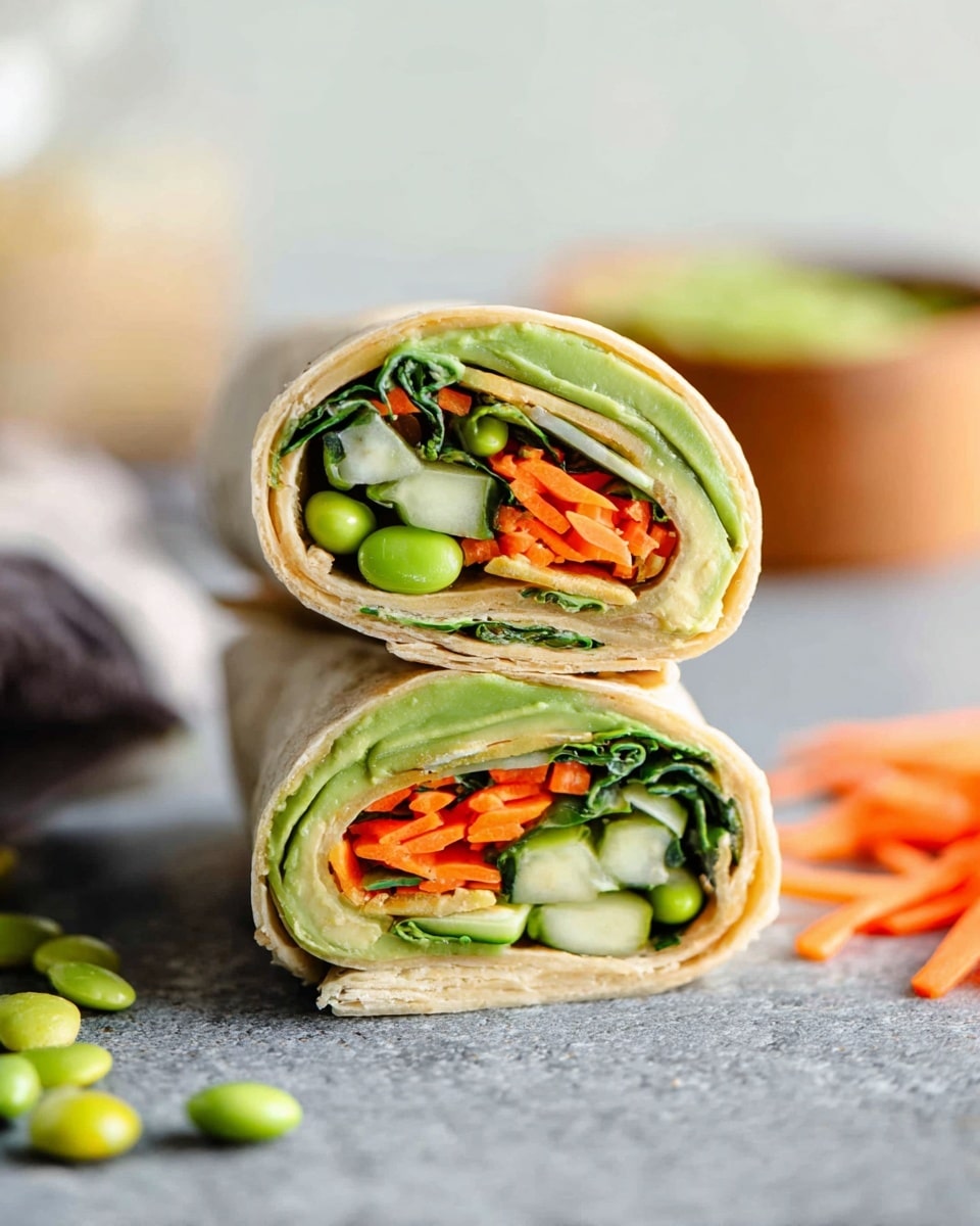 This image shows a close-up of a wrap cut in half and stacked vertically. The wrap has multiple visible layers: the outer layer is a light beige tortilla, inside which are bright green avocado slices with smooth texture, dark green leafy greens, thin orange carrot shreds, light green edamame beans, and pieces of cucumber with a fresh, pale-green color. The wrap sits on a gray surface with scattered carrot shreds and edamame. The background is softly blurred with light tones and hints of a bowl and other ingredients. photo taken with an iphone --ar 4:5 --v 7
