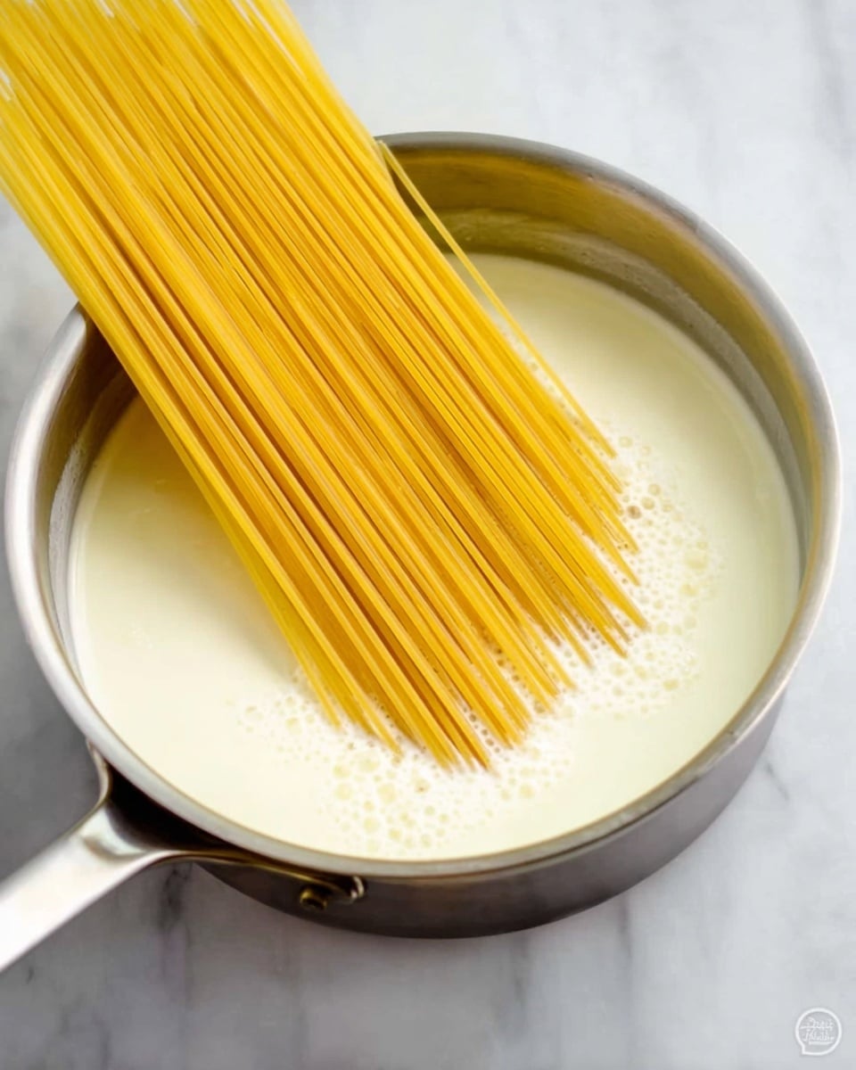 A silver pan filled with white creamy liquid, almost to the top. Over the pan, a bunch of long, straight, uncooked yellow spaghetti is placed diagonally from the bottom left corner to the top right, with one end touching the liquid and the other end resting on the pan’s edge. The background is a white marbled surface. photo taken with an iphone --ar 4:5 --v 7