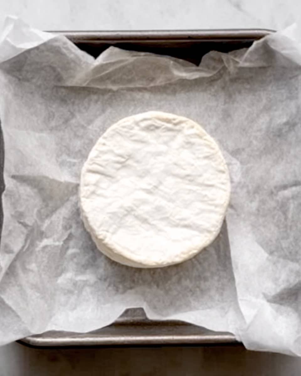 The image shows a round wheel of soft cheese with a white, powdery outer layer sitting on parchment paper inside a metal baking tray. The cheese has a smooth, creamy texture under the rind and is centered in the tray. The white parchment paper has some slight wrinkles around the cheese, adding a natural look. The tray is placed on a white marbled surface, giving a clean and bright background. Photo taken with an iphone --ar 4:5 --v 7