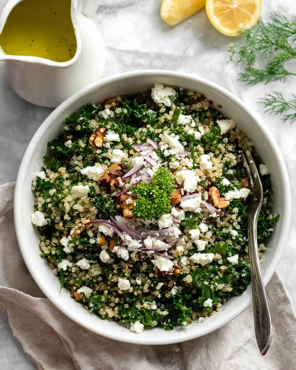 A deep white bowl filled with a fresh salad showing three main layers: the bottom layer is finely chopped kale with a dark green, leafy texture; the middle layer is light beige quinoa mixed evenly throughout; the top layer is small crumbles of white cheese scattered all over, with a few chopped brown nuts and thin strips of pale purple onion sprinkled on top. A small green herb sprig sits as garnish in the center. A silver fork rests inside the bowl, and a white sauce pitcher filled with yellow-green dressing is placed nearby on a white marbled surface, with a lemon wedge and small green herb sprigs partially visible in the background. photo taken with an iphone --ar 4:5 --v 7