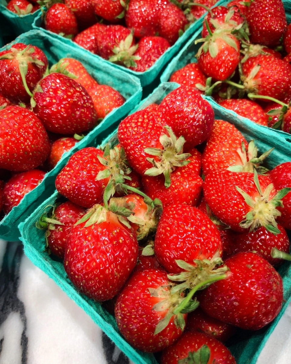 The image shows several green cardboard cartons filled with fresh, red strawberries. The strawberries are bright and shiny with some green leafy tops still attached. The cartons are tightly packed with the strawberries, showcasing their natural texture with small seeds visible on each berry. The background is a white marbled texture. photo taken with an iphone --ar 4:5 --v 7