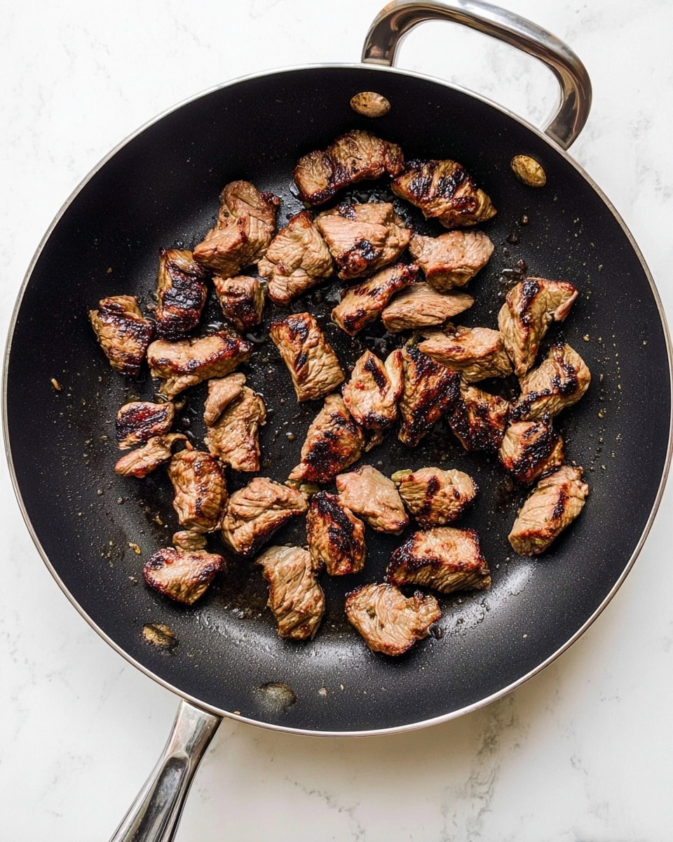 The image shows a black pan filled with small pieces of cooked meat. The meat pieces are browned and have dark grill marks, some parts look slightly crispy. They are spread across the pan’s surface with some spaces in between. The pan is placed on a white marbled textured surface, and the metal handle shines under the light. Photo taken with an iphone --ar 4:5 --v 7