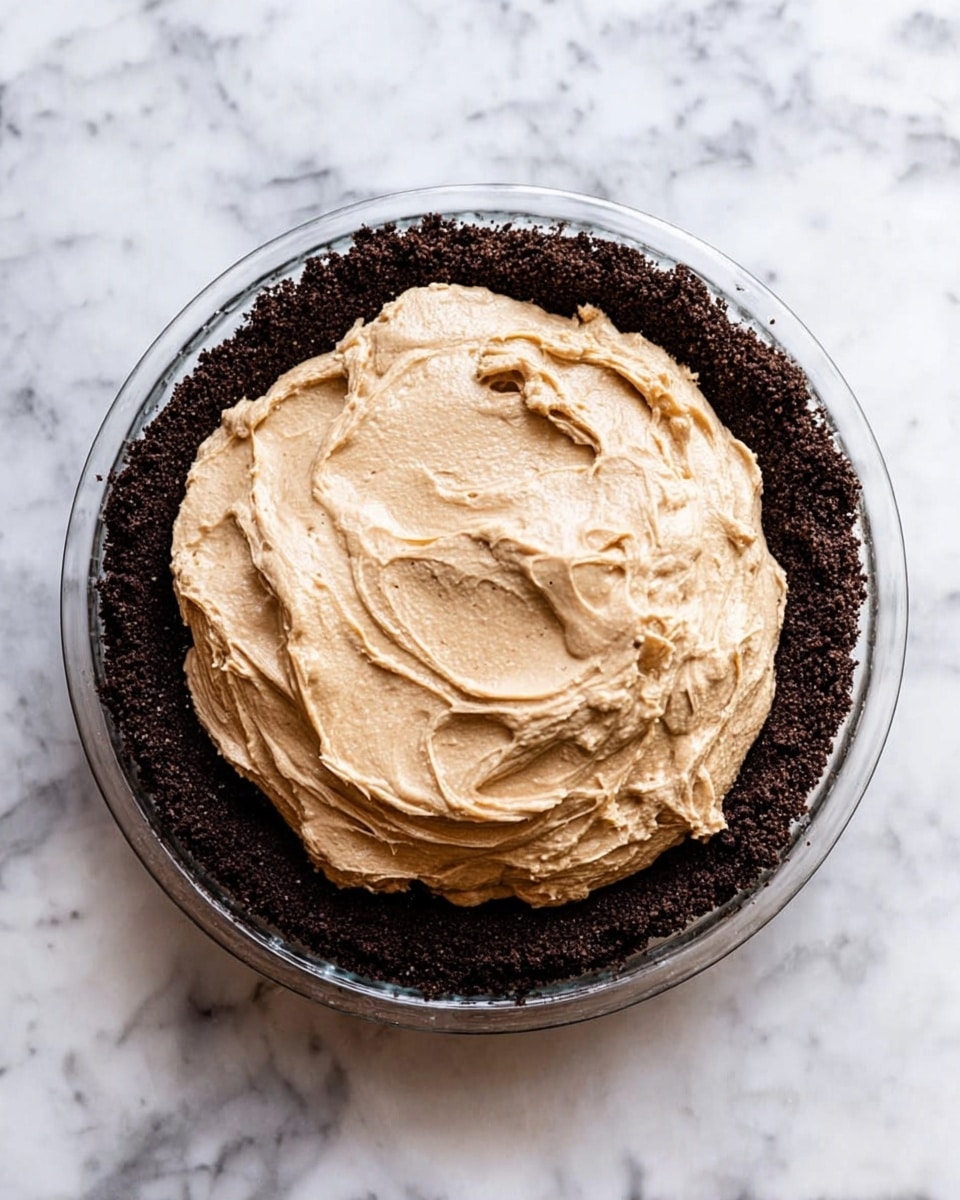A clear glass pie dish filled with two layers: the bottom layer is dark brown and crumbly, pressed evenly along the base and sides of the dish forming a crust; the top layer is a thick, creamy light brown filling piled high in the center with visible swirls and a smooth texture. The dish is placed on a white marbled surface. photo taken with an iphone --ar 4:5 --v 7