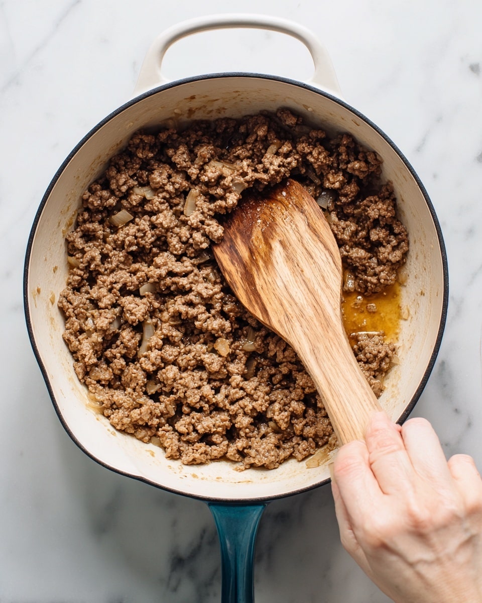 Inside a white enamel pot with a black rim, light brown cooked ground meat mixed with small bits of white onions covers the entire inside. A wooden spoon with visible grain is stirring the meat, held by a woman's hand matching the light skin tone. The pot is placed on a white marbled surface. The meat has a slightly chunky texture with some shiny oily liquid around. Photo taken with an iphone --ar 4:5 --v 7