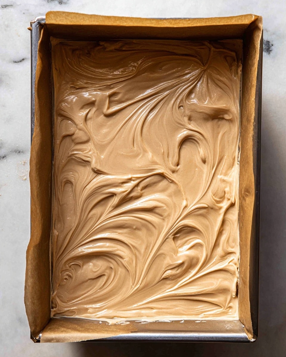 The image shows a single-layered dessert batter spread evenly inside a rectangular metal baking tray lined with brown parchment paper. The batter is smooth with swirled patterns, light brown in color, and creamy in texture with some soft peaks and swirls visible on the surface. The tray rests on a white marbled texture surface. photo taken with an iphone --ar 4:5 --v 7