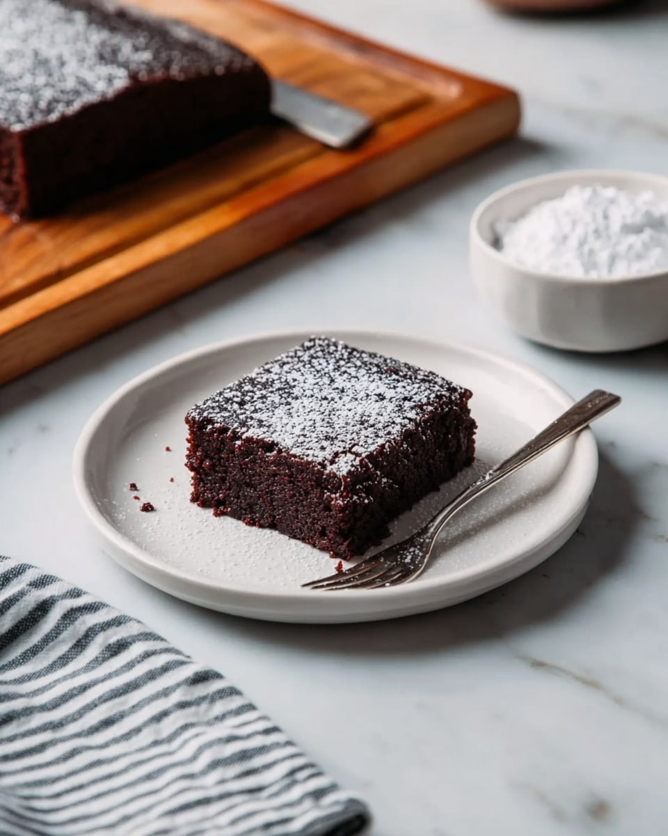 A square piece of dark brown chocolate cake sits in the center of a white plate, lightly dusted with white powdered sugar on top. The cake looks soft and dense with a slightly rough texture on its sides. A silver fork rests diagonally on the plate next to the cake. Behind the plate, there is a block of the same chocolate cake on a wooden board and a small white bowl filled with powdered sugar on a white marbled surface. A striped cloth napkin lies on the bottom left side of the image. photo taken with an iphone --ar 4:5 --v 7