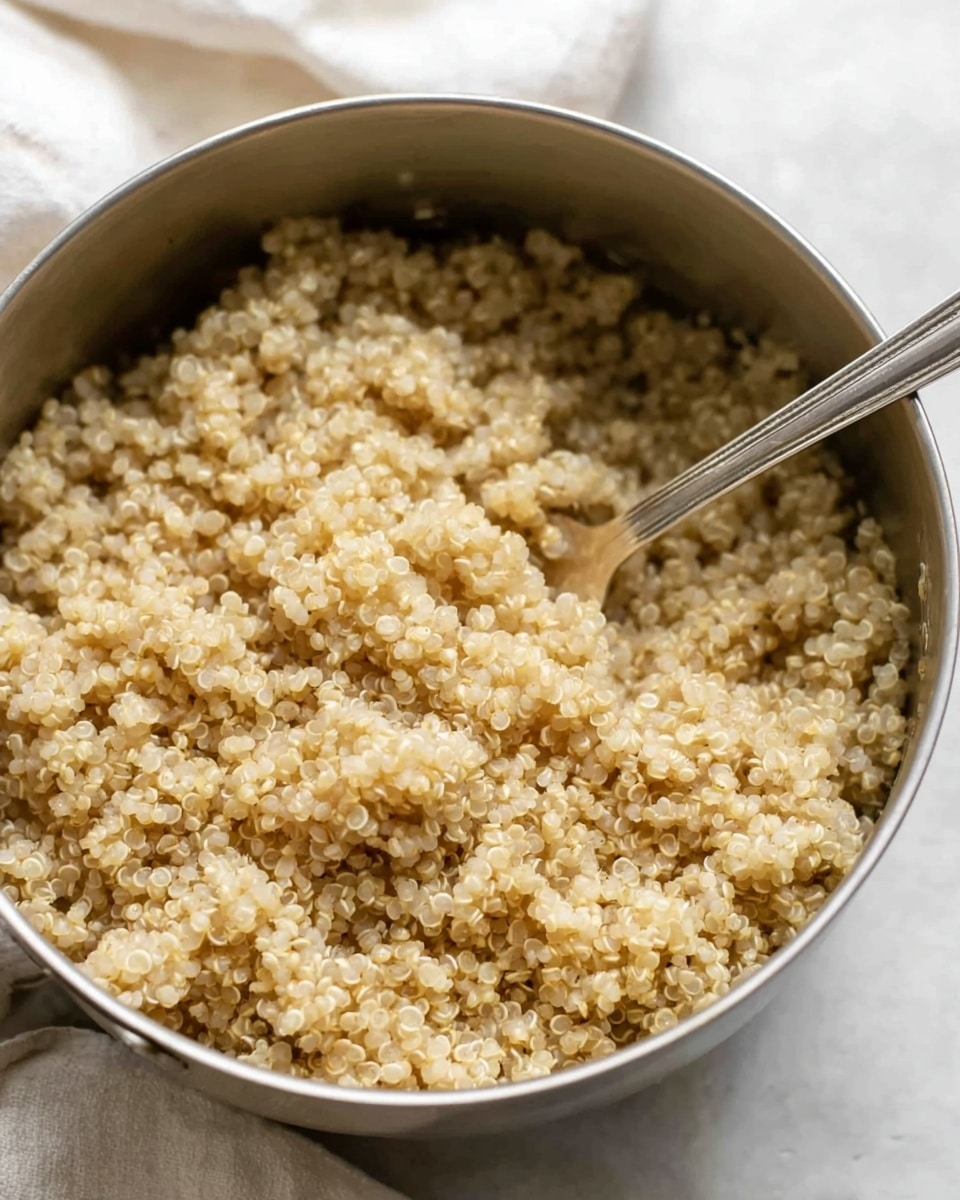 The image shows a close-up of a pot filled with cooked quinoa. The quinoa grains are small, round, and light beige with a slightly translucent look, creating a fluffy texture. A silver fork is partially buried in the quinoa, positioned towards the right side inside the stainless steel pot. The pot sits on a white marbled surface, and a white cloth is slightly visible in the background. Photo taken with an iphone --ar 4:5 --v 7