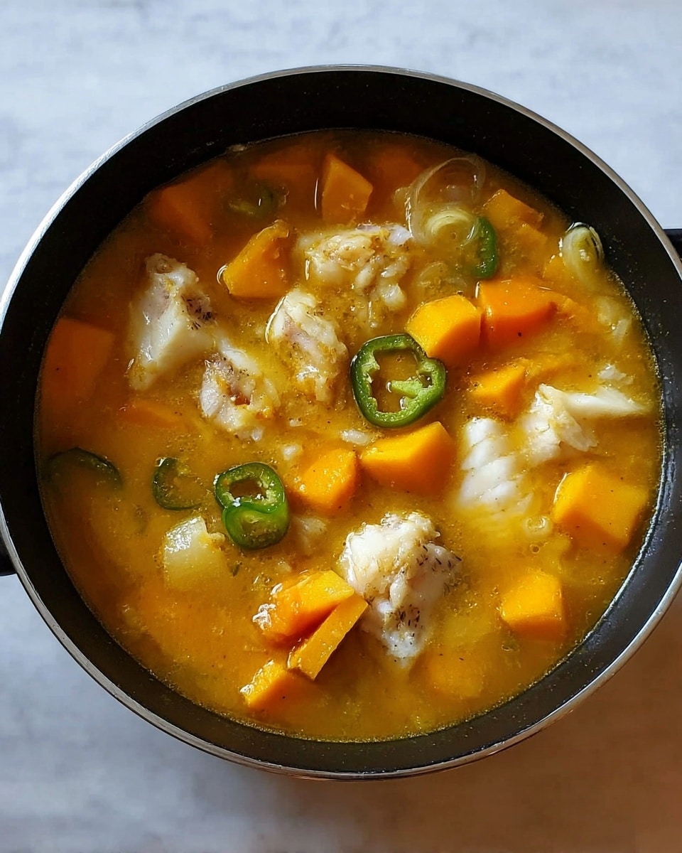 A black pan filled with a yellow-orange broth containing large chunks of white fish, bright orange cubes of squash, pieces of onion, and green slices of peppers. The soup looks thick and hearty, with the ingredients floating evenly throughout the pan. The background is a white marbled surface. photo taken with an iphone --ar 4:5 --v 7