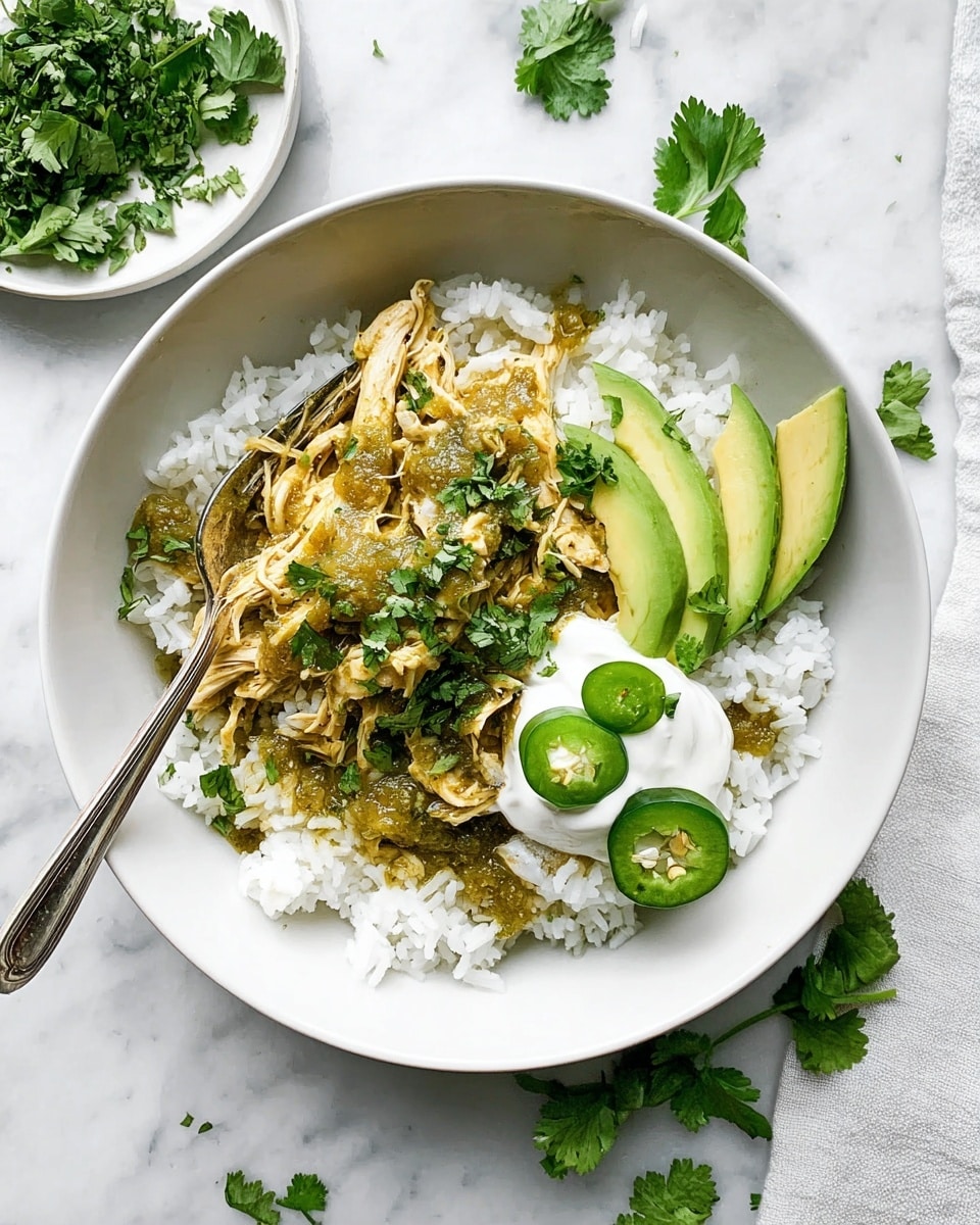 A white bowl sits on a white marbled surface, filled with a base layer of fluffy white rice. On top, shredded chicken is covered in green sauce, sprinkled with chopped fresh cilantro leaves. To the side of the chicken, two slices of green avocado rest next to a dollop of white sour cream, topped with three slices of green jalapeño peppers and a few cilantro leaves. A silver fork is placed inside the bowl on the left side. In the background, some fresh cilantro leaves are spread on a small white plate and also scattered on the white marbled surface. photo taken with an iphone --ar 4:5 --v 7