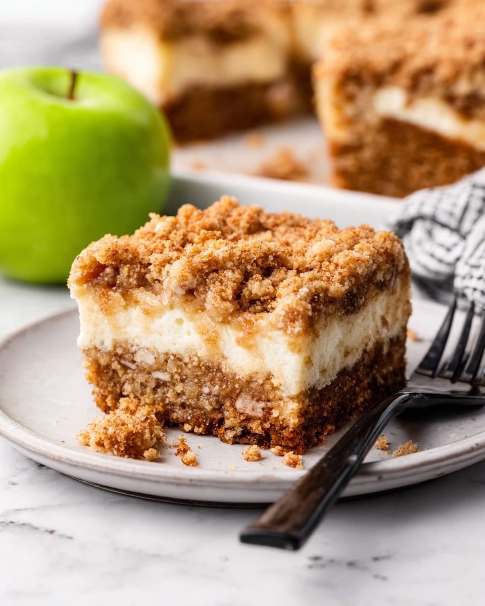 A white plate holds a square serving of crumb cake with a golden crumbly top layer, a thick creamy white middle layer, and a dense brown bottom layer filled with small nut pieces. The crumb topping appears rough and crunchy. A shiny black fork with a piece of the crumb cake is placed on the plate. In the background, there is a green apple and another plate with the crumb cake, all set on a white marbled surface. photo taken with an iphone --ar 4:5 --v 7