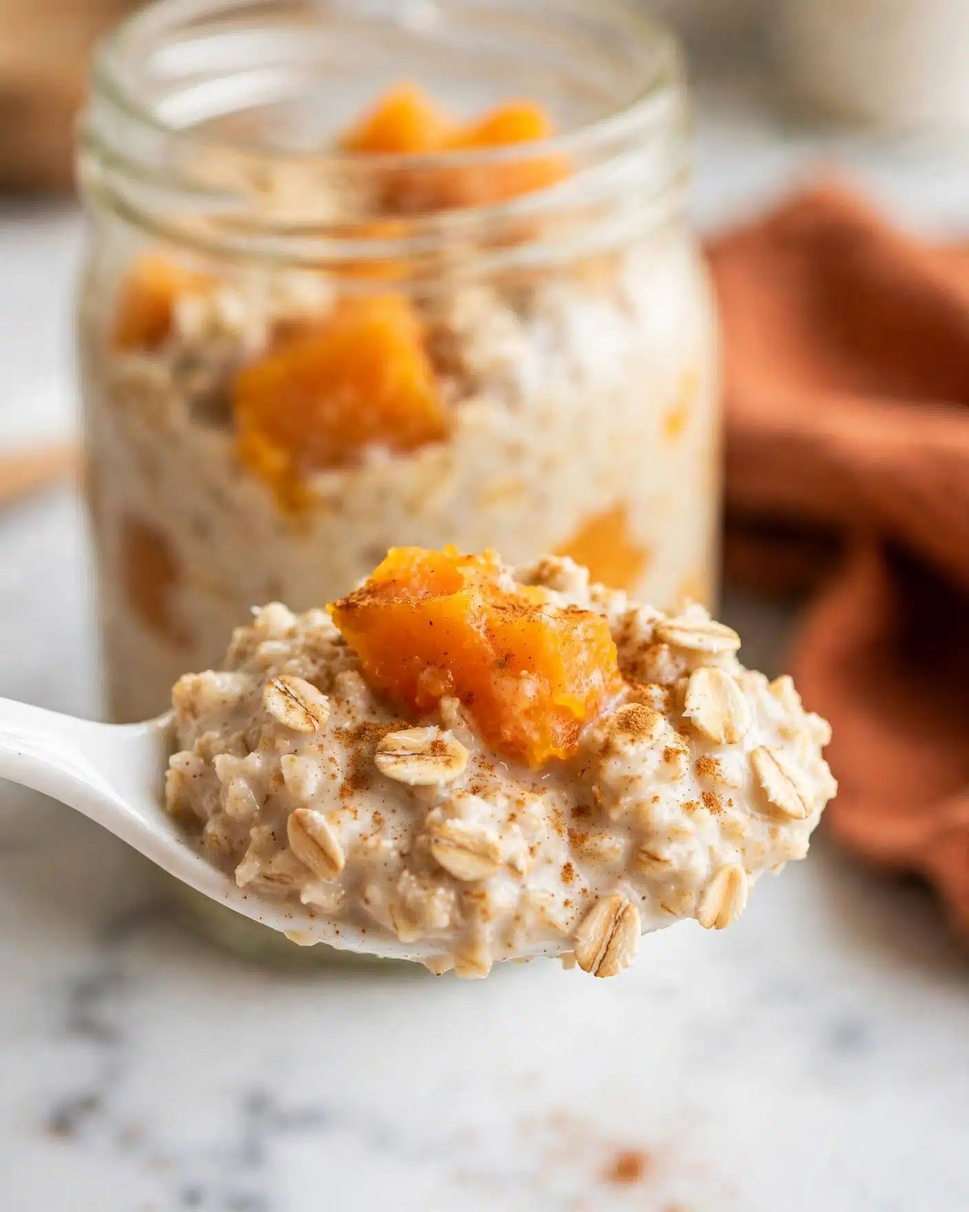 A close-up of a white spoon holding a creamy oatmeal mixture with visible oats and a bright orange piece of cooked fruit, possibly pumpkin or sweet potato, dusted lightly with cinnamon or spice. In the blurred background, there is a clear glass jar filled with the same oatmeal and fruit blend, placed on a white marbled surface. The colors are warm, with soft orange hues contrasting against the creamy oatmeal texture. A soft focus shows part of a rust-colored cloth to the side. The scene is bright with natural lighting, showing a cozy and fresh atmosphere. Photo taken with an iphone --ar 4:5 --v 7