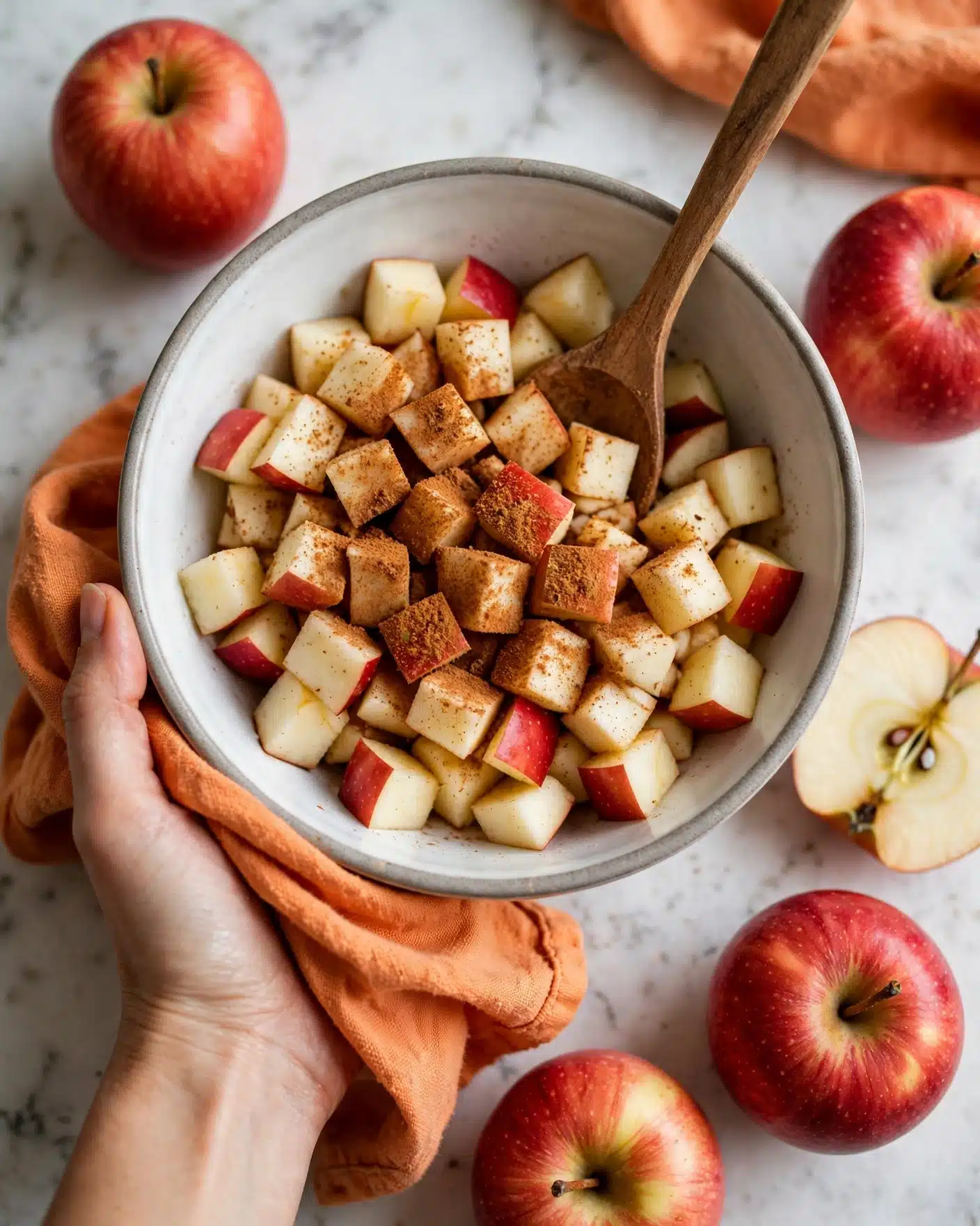 A white bowl filled with pieces of red and white apple chunks mixed with brown cinnamon powder on top, showing a rough texture of the spice dusting the fruit evenly, held by a woman's hand wrapped in a soft orange cloth at the bottom left, while another woman's hand stirs the mixture with a small wooden spoon from the upper right side; the background is a white marbled surface with an apple cut in half and a whole apple on the top left side, adding a fresh and cozy atmosphere. photo taken with an iphone --ar 4:5 --v 7