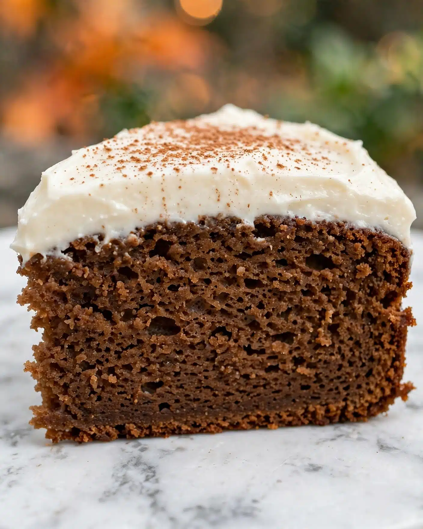Pumpkin Dream Cake Recipe 5 A single slice of thick brown cake with a soft, moist texture is shown in close-up. The cake has a dense crumb with small holes inside. It is topped with a thick, creamy white frosting layer that looks smooth and rich, sprinkled lightly with a fine brown powder on top. The background is a soft blur with hints of orange and green, and the cake sits on a surface with a white marbled texture. photo taken with an iphone --ar 4:5 --v 7