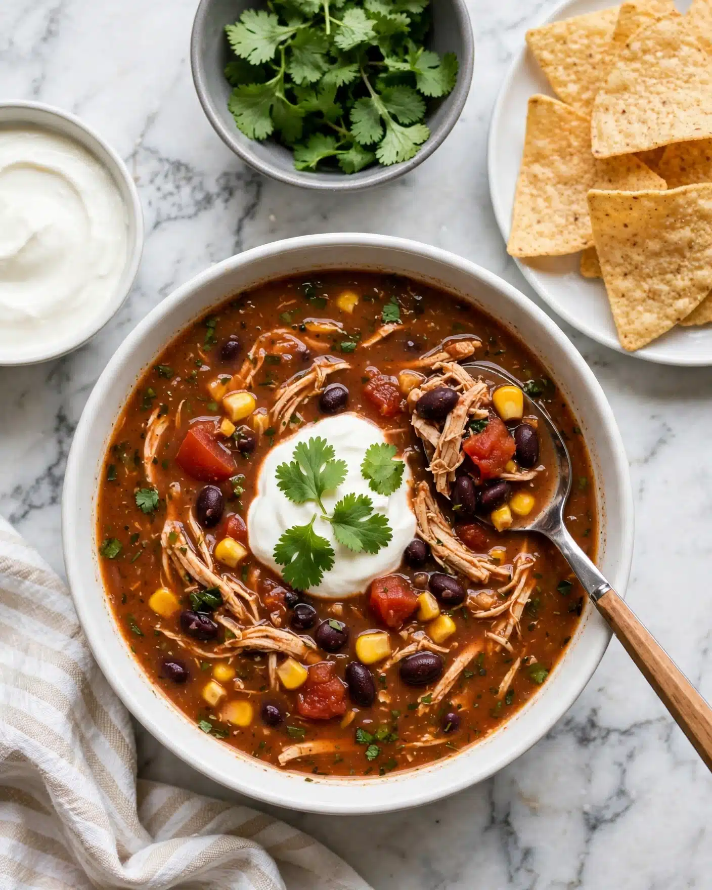 My Favorite Slow Cooker Chicken Chili Recipe 5 A white bowl filled with thick reddish-brown soup containing visible layers of shredded chicken, black beans, diced tomatoes, yellow corn, and small green herbs mixed throughout. The soup has a dollop of white sour cream topped with fresh green cilantro leaves in the center. A silver spoon with a wooden handle rests inside the bowl on the right side, scooping some soup. To the top right, a white plate holds several light golden tortilla chips with a rough texture. Above the bowl, a small gray bowl contains fresh bright green cilantro leaves. To the left, there is a small white bowl filled with creamy white sour cream. All items rest on a white marbled surface with a soft striped kitchen towel partially visible at the bottom left corner. Photo taken with an iphone --ar 4:5 --v 7