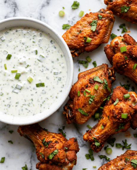 The image shows a close-up of a white bowl filled with creamy white dipping sauce speckled with small green onion pieces on the left. Surrounding the bowl are several golden brown chicken wings with a crispy, spicy coating in shades of orange and reddish brown. The wings are sprinkled with small pieces of chopped green onions and cilantro, adding pops of green color. The white marbled surface beneath adds a clean, bright background that contrasts with the warm colors of the chicken. photo taken with an iphone --ar 4:5 --v 7