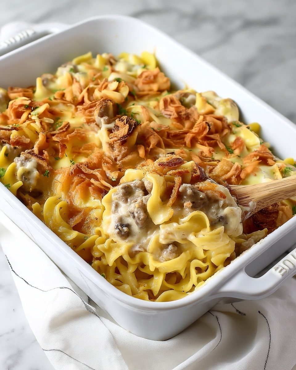 A white rectangular baking dish filled with a creamy pasta casserole, showing layers of wide, curly noodles at the bottom, covered by a thick white sauce with visible pieces of ground meat and small green herbs mixed in; on top of the sauce are golden crispy fried onions scattered evenly across the surface, adding texture and color contrast to the dish. A wooden spoon lifts a portion of the casserole, displaying a close-up of the creamy sauce with ground meat and crispy onions on top, against a white marbled surface background. Photo taken with an iphone --ar 4:5 --v 7