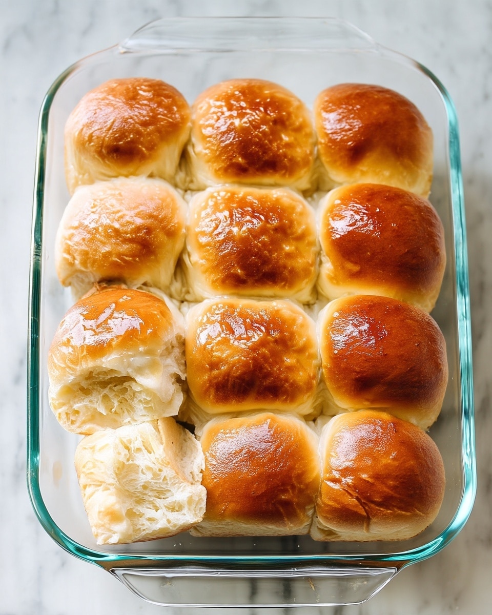 A close-up view of a glass baking dish filled with nine golden brown dinner rolls arranged in three rows of three. Each roll has a shiny, slightly textured top with light browning. A brush with light wood handle and metallic bristles is brushing melted butter on the top of the front-left roll, creating a glistening, smooth, wet layer on its surface. The baking dish sits on a white marbled surface, adding a clean and bright background to the image. The photo taken with an iphone --ar 4:5 --v 7