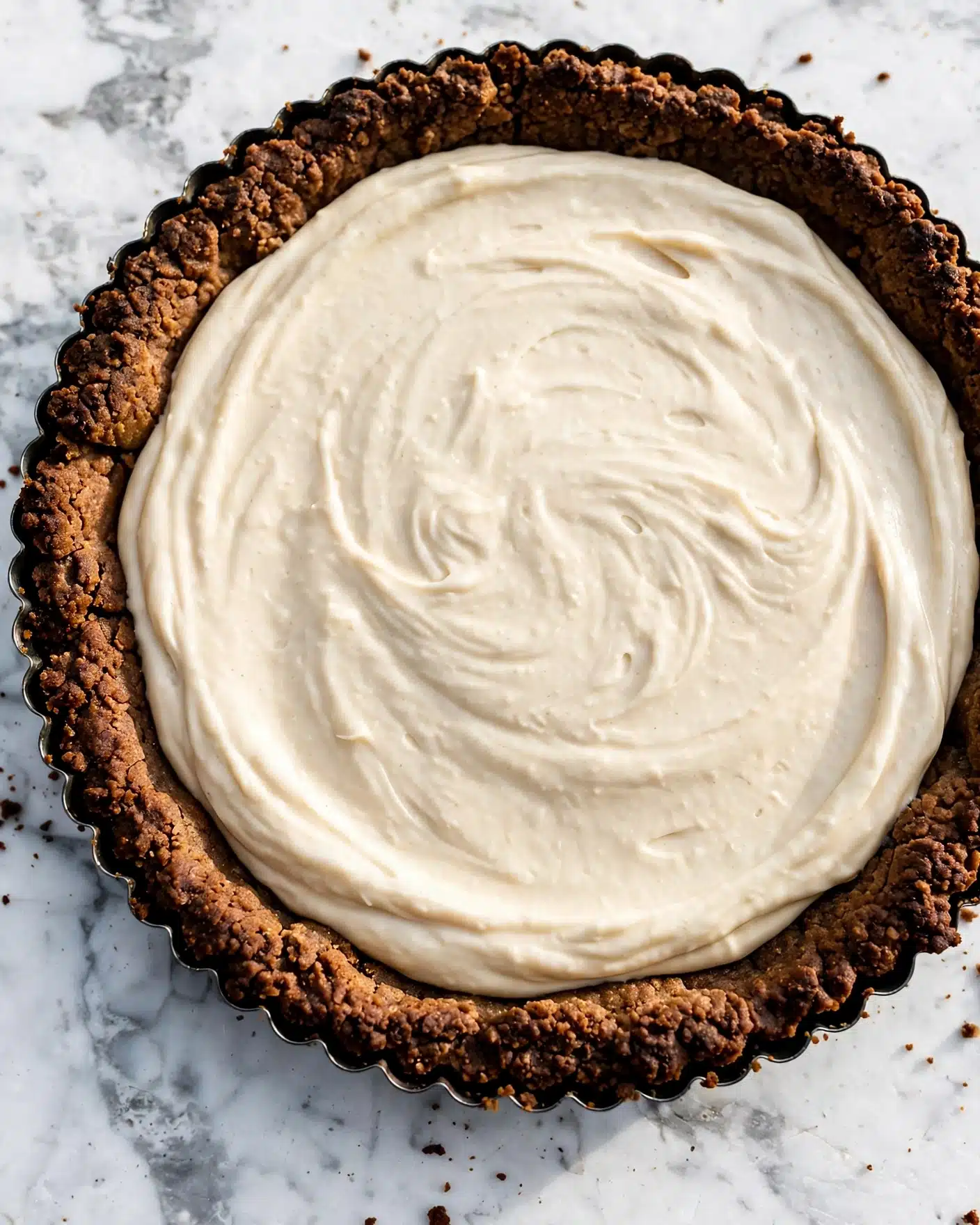 A tart with two visible layers: the bottom layer is a rough, dark brown crust that looks crunchy and thick, filling the fluted edges of the round tart pan, and the top layer is a smooth, creamy light beige filling spread evenly inside the crust, with gentle swirls on the surface. The tart is placed on a white marbled textured surface. photo taken with an iphone --ar 4:5 --v 7