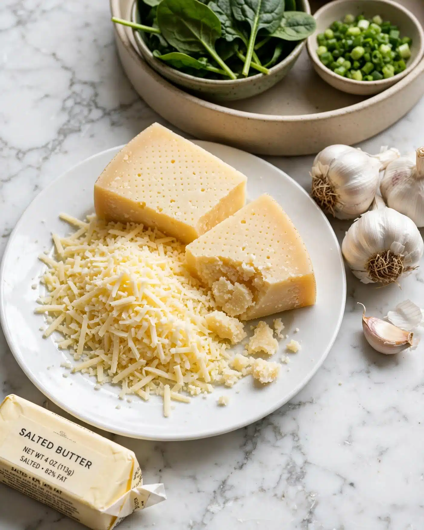 A close-up of a white plate filled with finely grated pale yellow hard cheese, with two wedge pieces of the same cheese resting on top, showing dotted patterns on their surfaces and a crumbly texture where one wedge is broken. Next to the plate, there are three whole garlic bulbs and one separated clove with a papery white skin. Behind them, there is a beige dish holding a small green bowl filled with fresh dark green spinach leaves and another small bowl with chopped green onions. On the bottom left corner of the image, there is a wrapped package of salted butter in pale yellow paper with black writing. The scene is set on a white marbled textured surface. Photo taken with an iphone --ar 4:5 --v 7