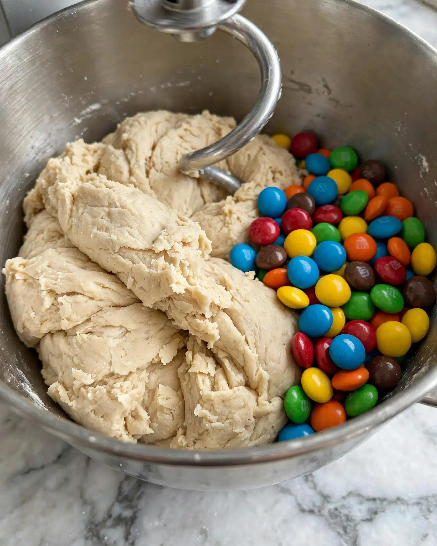 Monster Cookies Recipe 4 A close-up view of a silver mixing bowl with dough and colorful candy-coated chocolates sitting together inside. The dough is light beige, soft, and thick, taking up about half the bowl, with folded and slightly rough texture. The bright candies in blue, red, yellow, green, orange, and brown form a vibrant pile to one side, showing smooth surfaces and round shapes. A silver mixer attachment partly dips into the dough from above. The bowl is placed on a white marbled surface. photo taken with an iphone --ar 4:5 --v 7