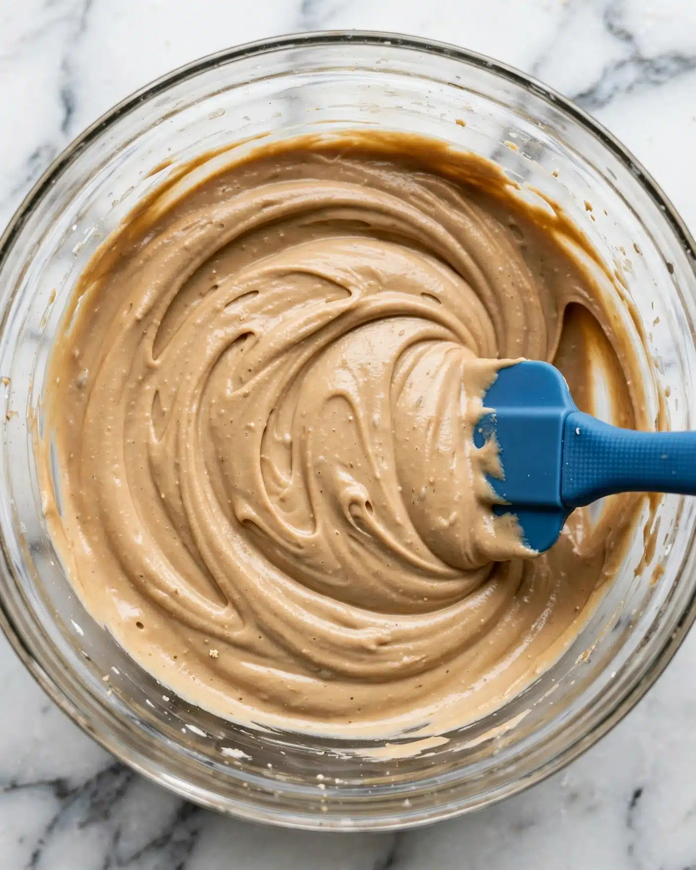 A close-up view of a clear glass bowl filled with light brown creamy mixture that has smooth, thick, and swirled texture spread evenly around the bowl, with a blue silicone spatula partly submerged on the right side, all set on a white marbled surface photo taken with an iphone --ar 4:5 --v 7