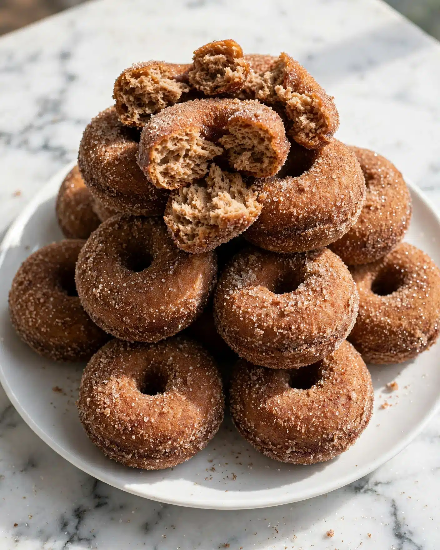 Baked Apple Cider Donuts Recipe 5 A white plate is filled with a stack of about ten small round donuts, each coated in a layer of sugar and cinnamon. The donuts have a rough, slightly crunchy brown surface with a central hole and look soft inside. On the top of the pile, one donut is broken into pieces showing a light brown, soft texture, while the others keep their full round shape. The plate sits on a white marbled surface, and the lighting highlights the sugar crystals on the donuts. photo taken with an iphone --ar 4:5 --v 7