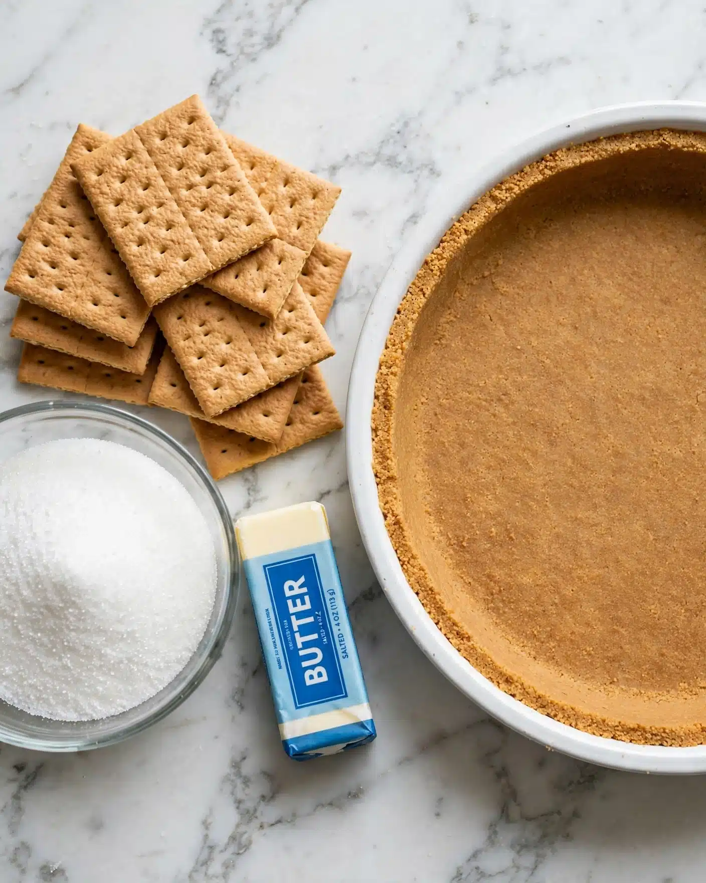 Pecan Pie Cheesecake Recipe 4 The image shows two parts side by side on a white marbled surface. On the left side, there are several whole square graham crackers stacked unevenly, with a clear glass bowl filled with white granulated sugar below them, and a stick of butter with a blue label laying horizontally to the right of the bowl. On the right side, there is a white round pan filled with a smooth, evenly pressed single layer of golden-brown graham cracker crust covering the bottom and sides evenly. The photo taken with an iphone --ar 4:5 --v 7