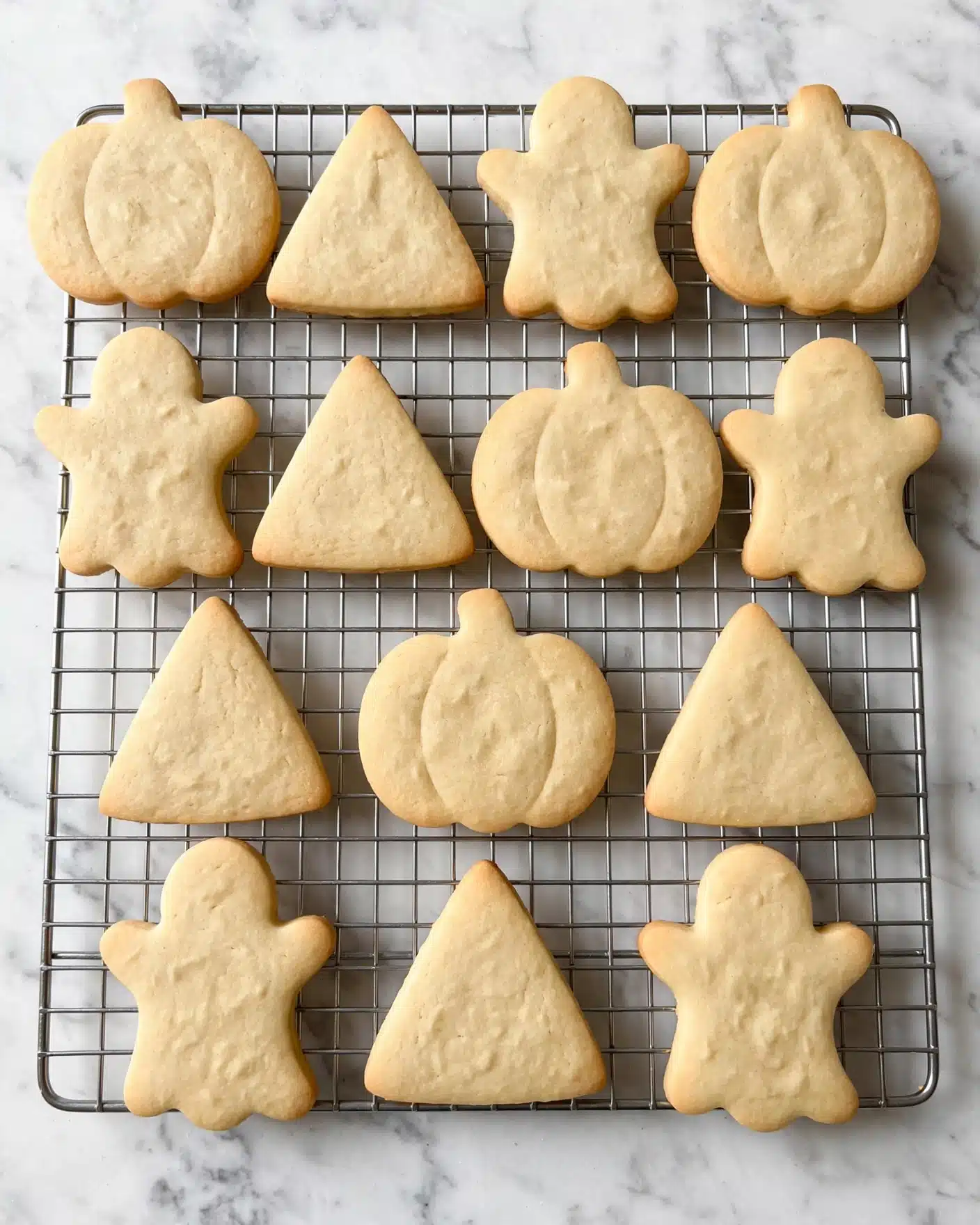 The image shows fourteen plain sugar cookies shaped like pumpkins, ghosts, and triangles on a silver cooling rack. The cookies are light golden in color with a smooth texture and soft edges. The cooling rack is placed on a white marbled surface, providing a clean and simple background. The cookies are evenly spaced, showing their different shapes clearly and with no decoration on them photo taken with an iphone --ar 4:5 --v 7