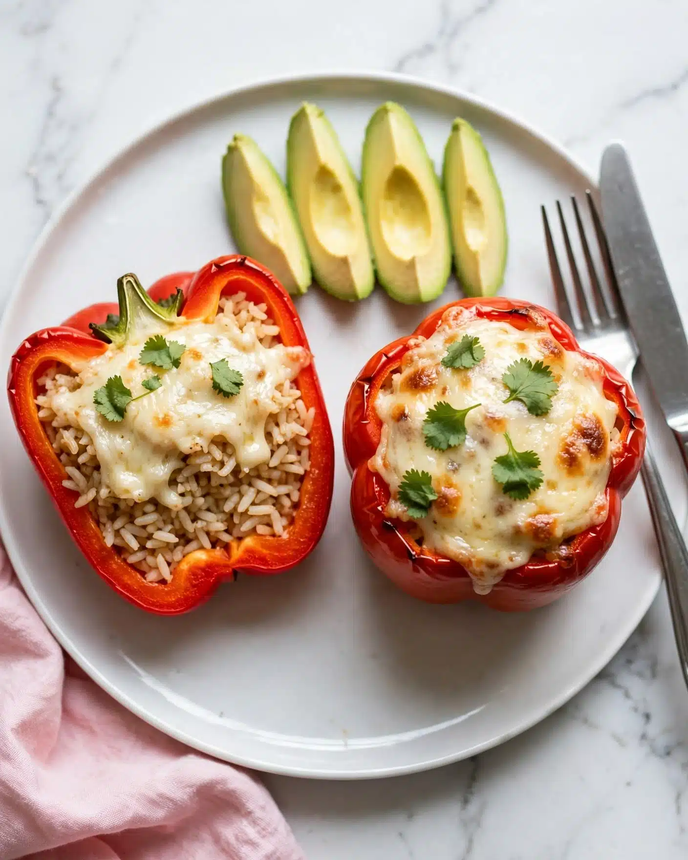 A white plate with a stuffed red bell pepper cut in half on the left side, showing layers of cooked rice and melted white cheese on top, garnished with small green cilantro leaves. On the right side of the plate, there is a whole stuffed red bell pepper with melted cheese and cilantro on top. Above the peppers, there are four slices of yellow avocado with green edges. A silver knife and fork rest on the right side of the plate. The plate is on a white marbled background, with a pink cloth partially visible at the bottom left corner. Photo taken with an iphone --ar 4:5 --v 7