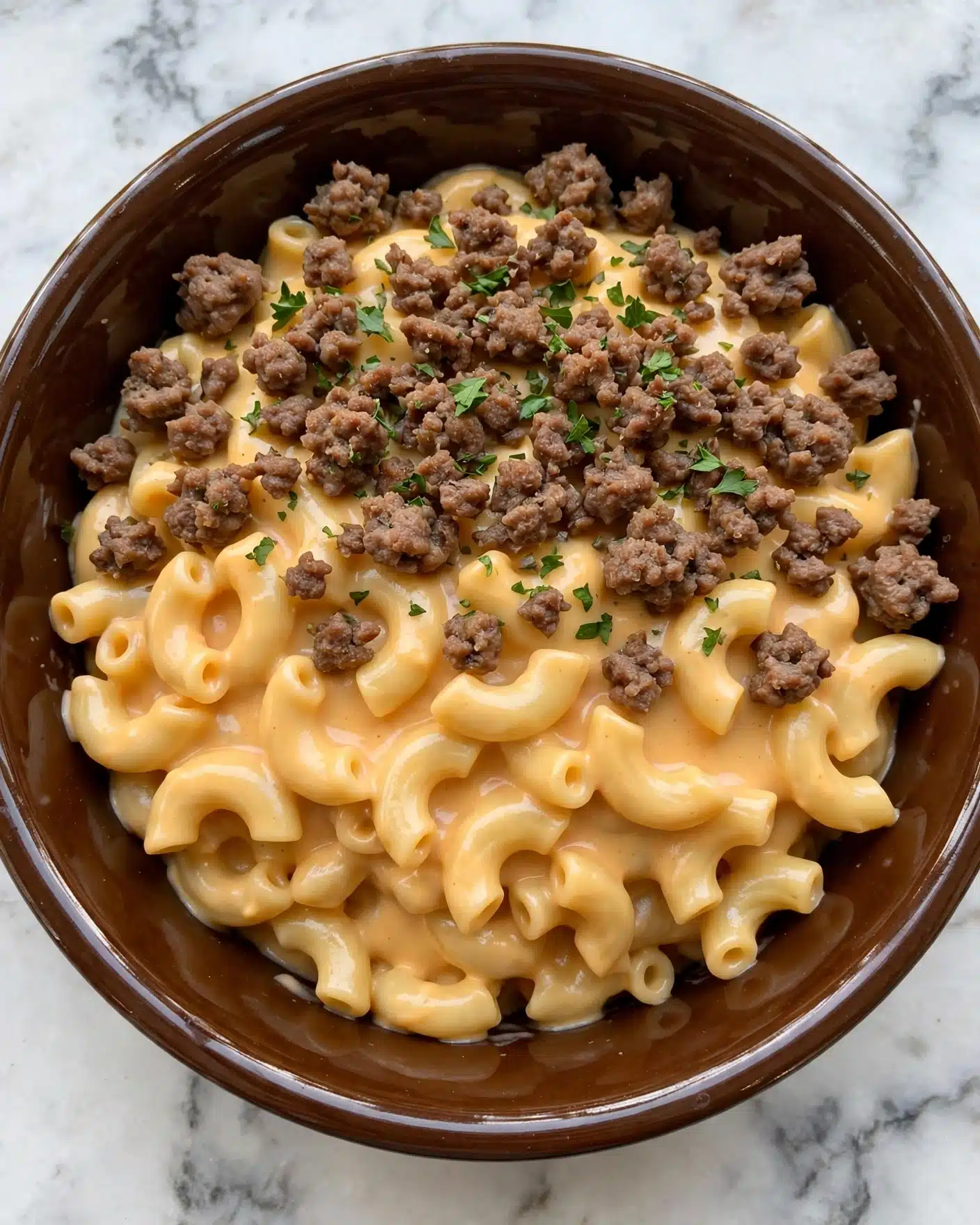 A close-up view of creamy macaroni and ground meat in a shiny brown bowl, showing about two layers mixed together; the first layer features tender elbow macaroni pasta coated in a smooth, light orange cheese sauce with a soft texture, while the second layer has small, browned chunks of ground meat scattered evenly throughout, topped with tiny green parsley flakes for color. The bowl sits on a white marbled surface. Photo taken with an iphone --ar 4:5 --v 7